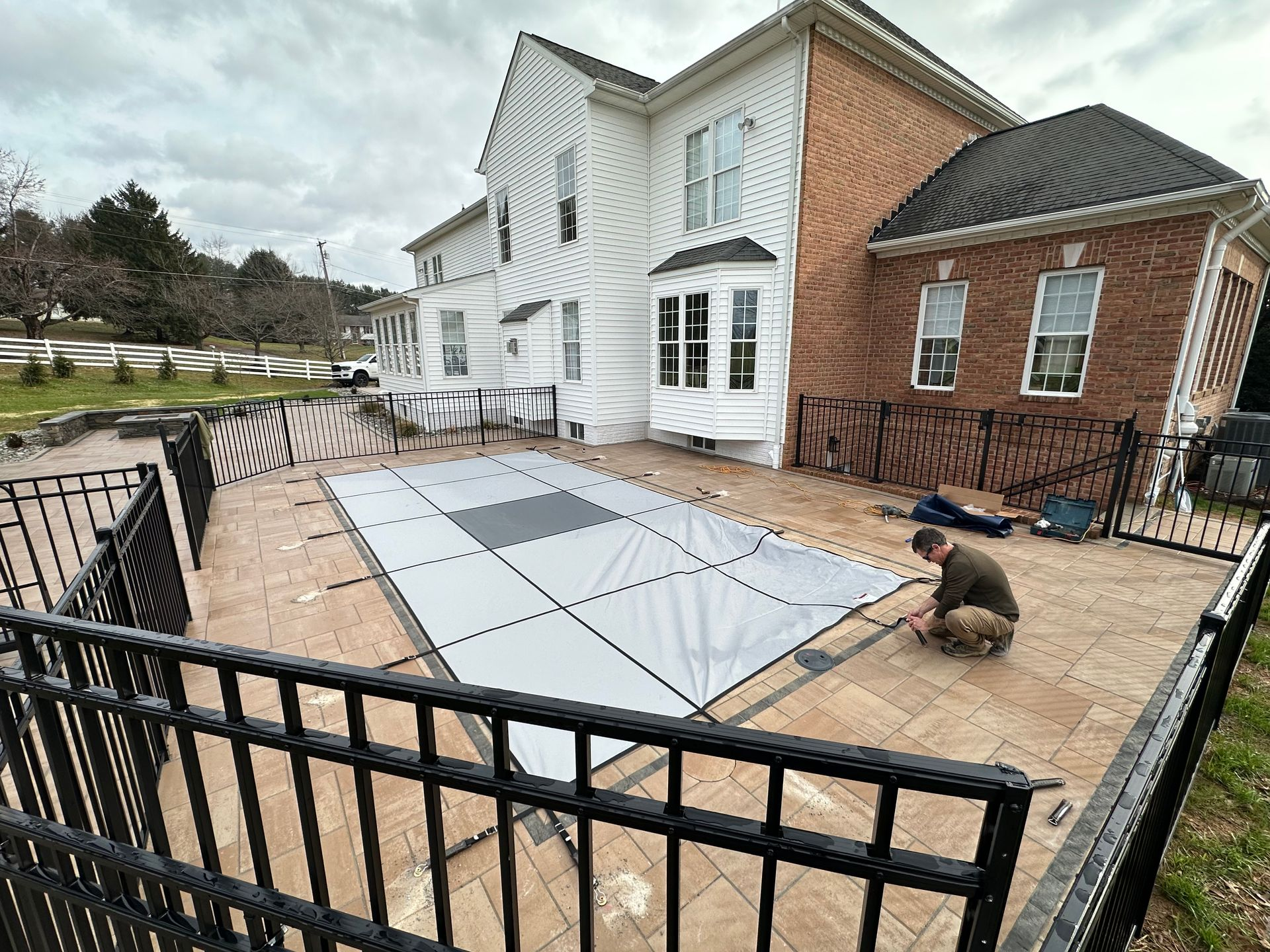 Pool cover being installed on a patio, with a house in the background. A person kneels, working.