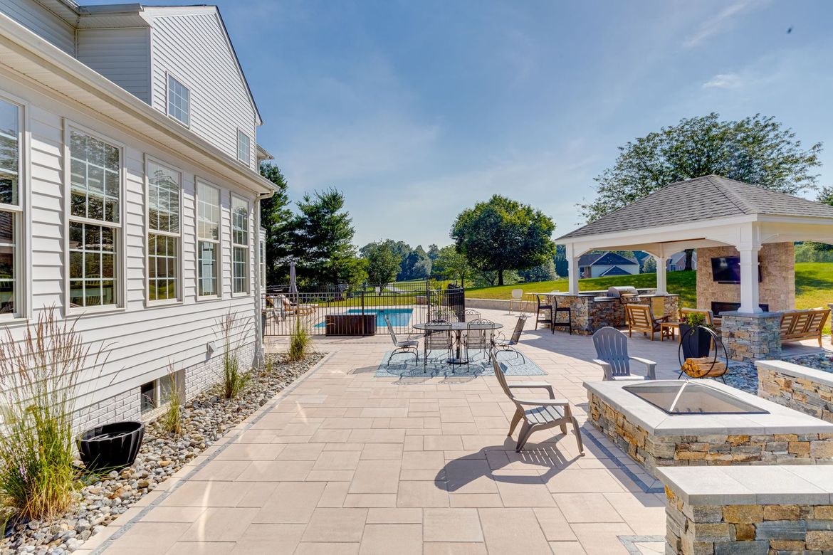 Patio with seating, fireplace, and gazebo beside a pool and house under a sunny sky.