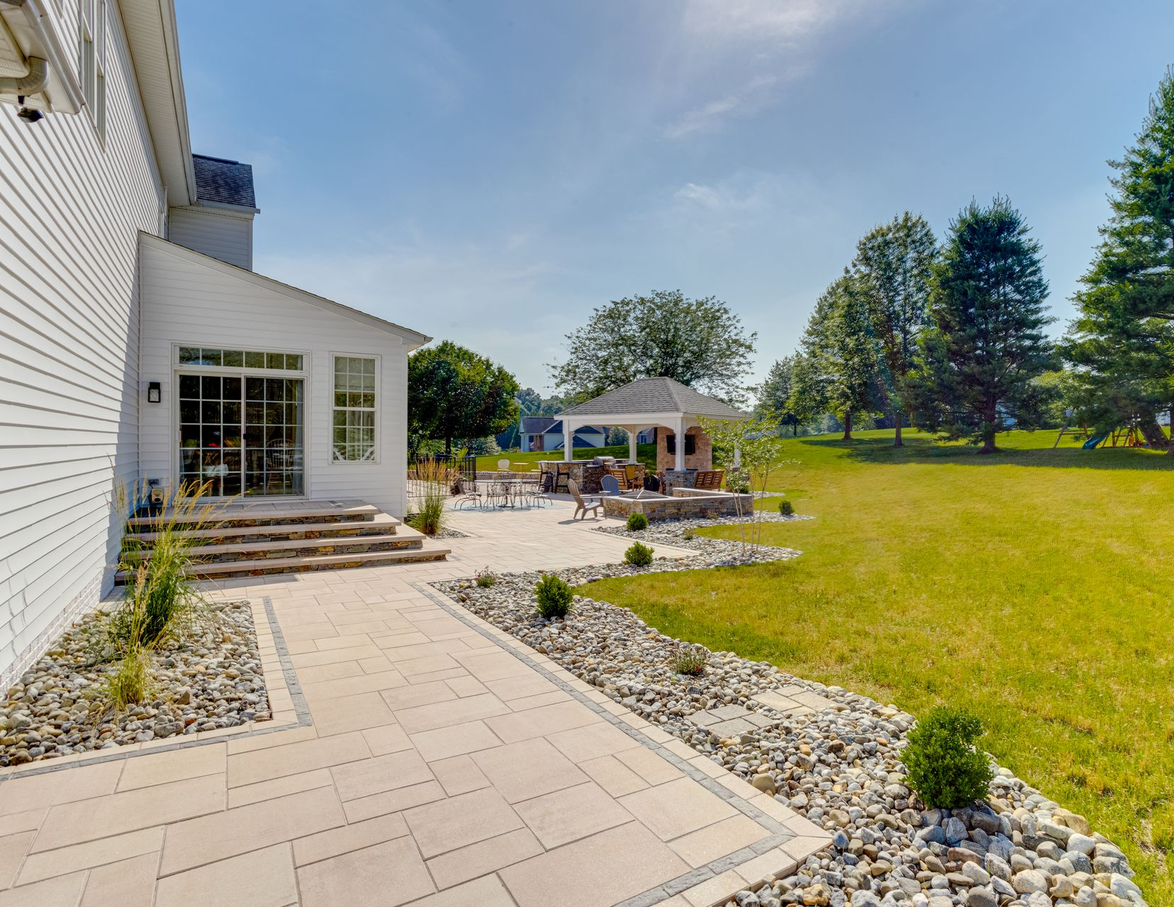 A paved walkway leads to a white house patio with a gazebo and large backyard on a sunny day.