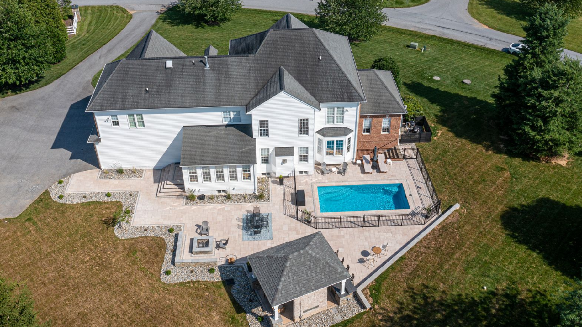 Aerial view of a white multi-story house with a backyard pool, stone patio, and a covered pavilion on a sunny day.