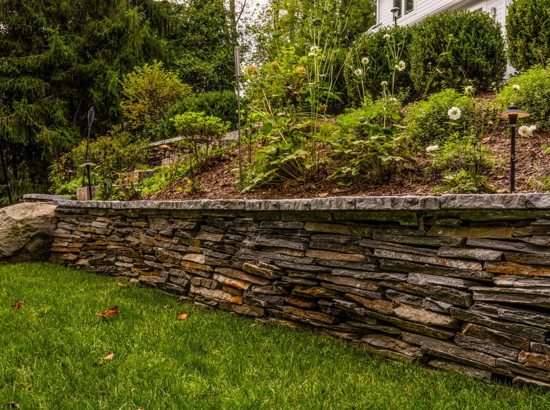 Stone retaining wall supporting a hillside garden with green grass and bushes.