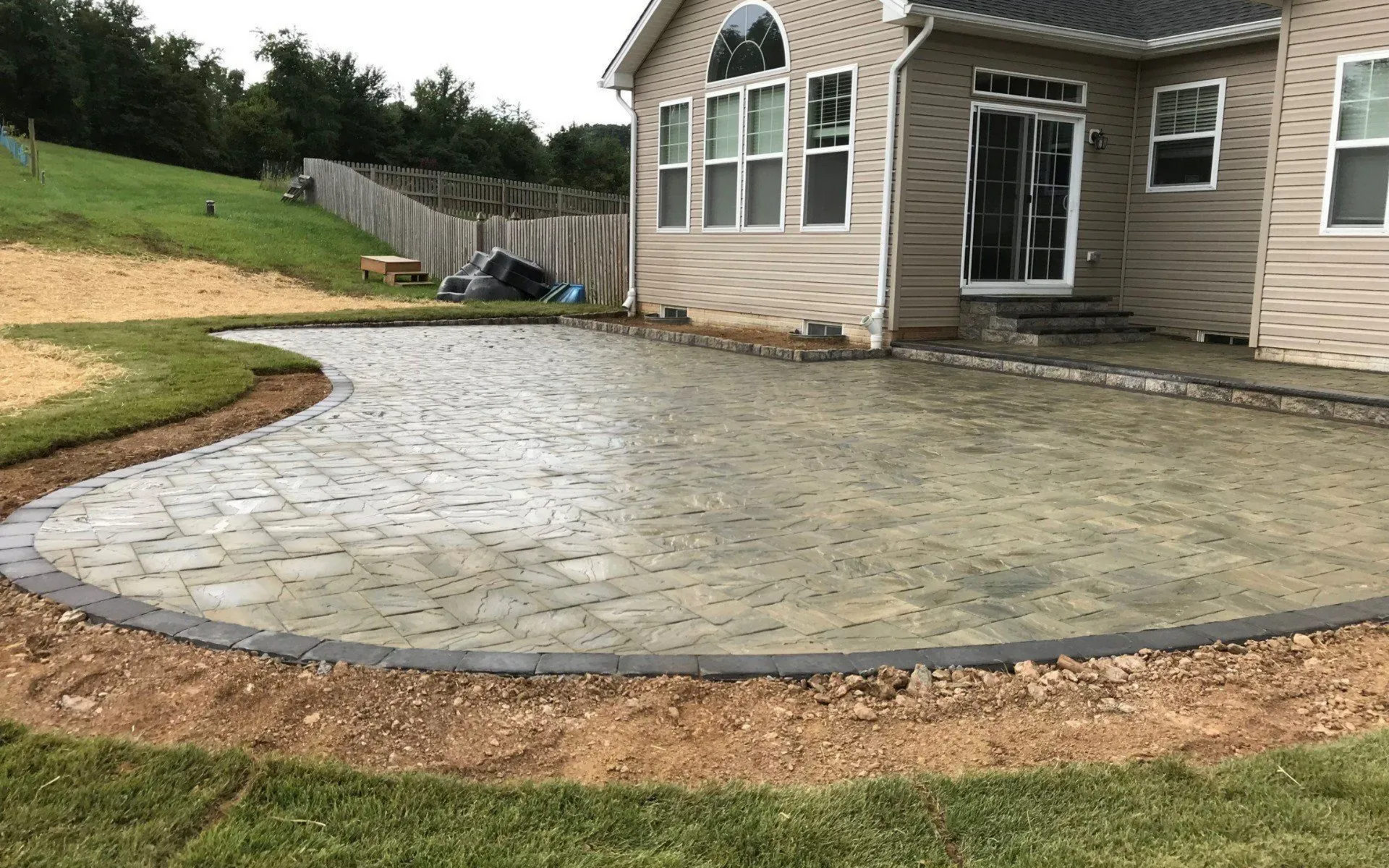 A new stone patio next to a house with grass and a hillside in the background.