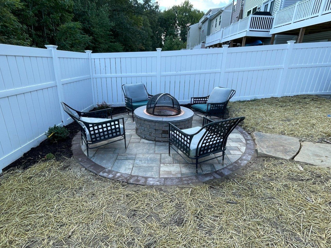 A fire pit is surrounded by chairs and a white fence in a backyard.