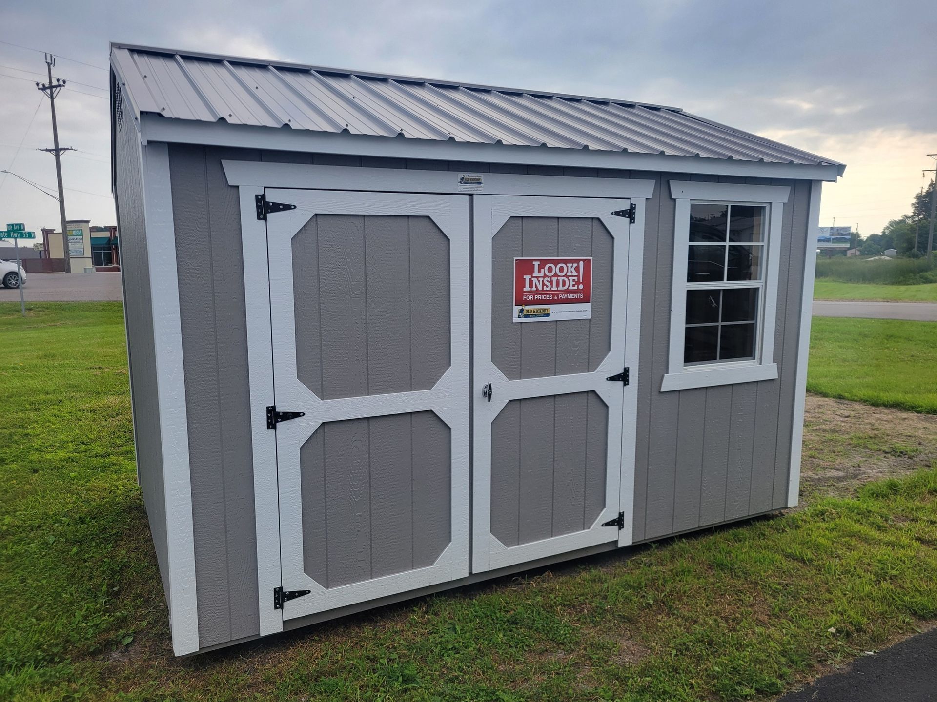 Gray shed with white trim, double doors, metal roof, and a window on a grassy area.
