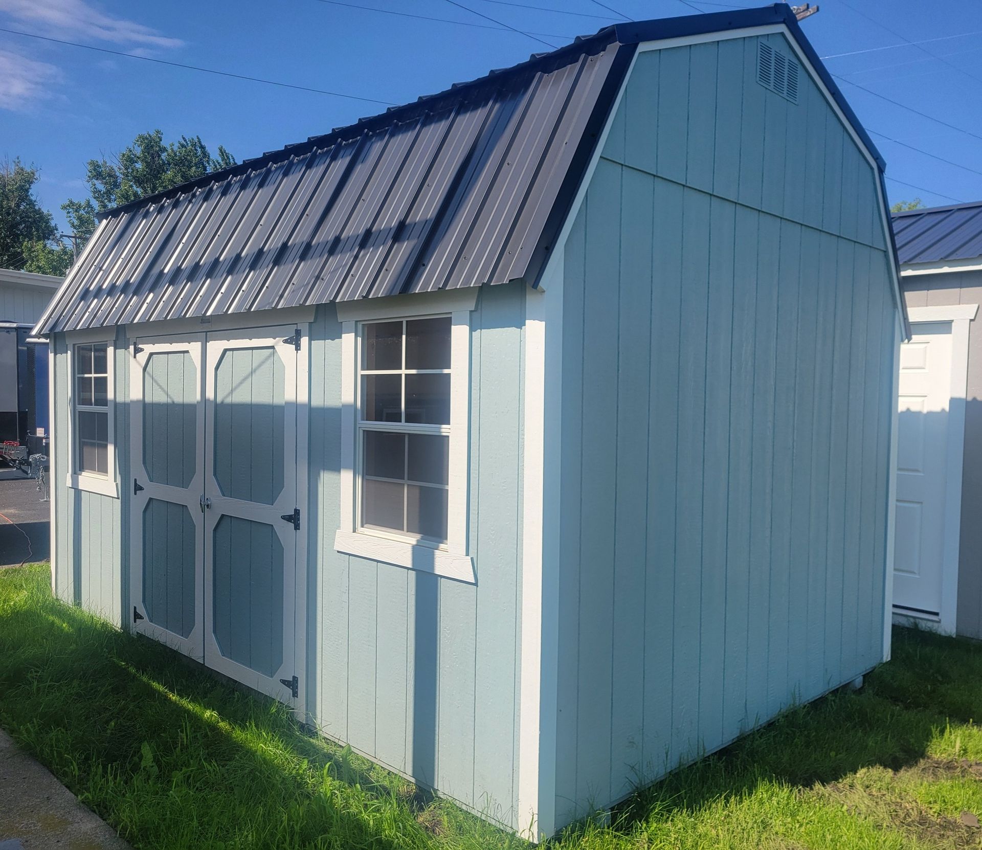 Light blue shed with black roof, white trim, and double doors, set on green grass.