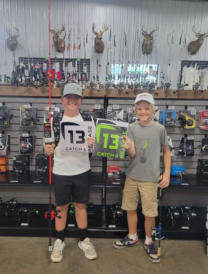 Two boys holding fishing gear in a store, one with a number 13, smiling. Fishing equipment on shelves.