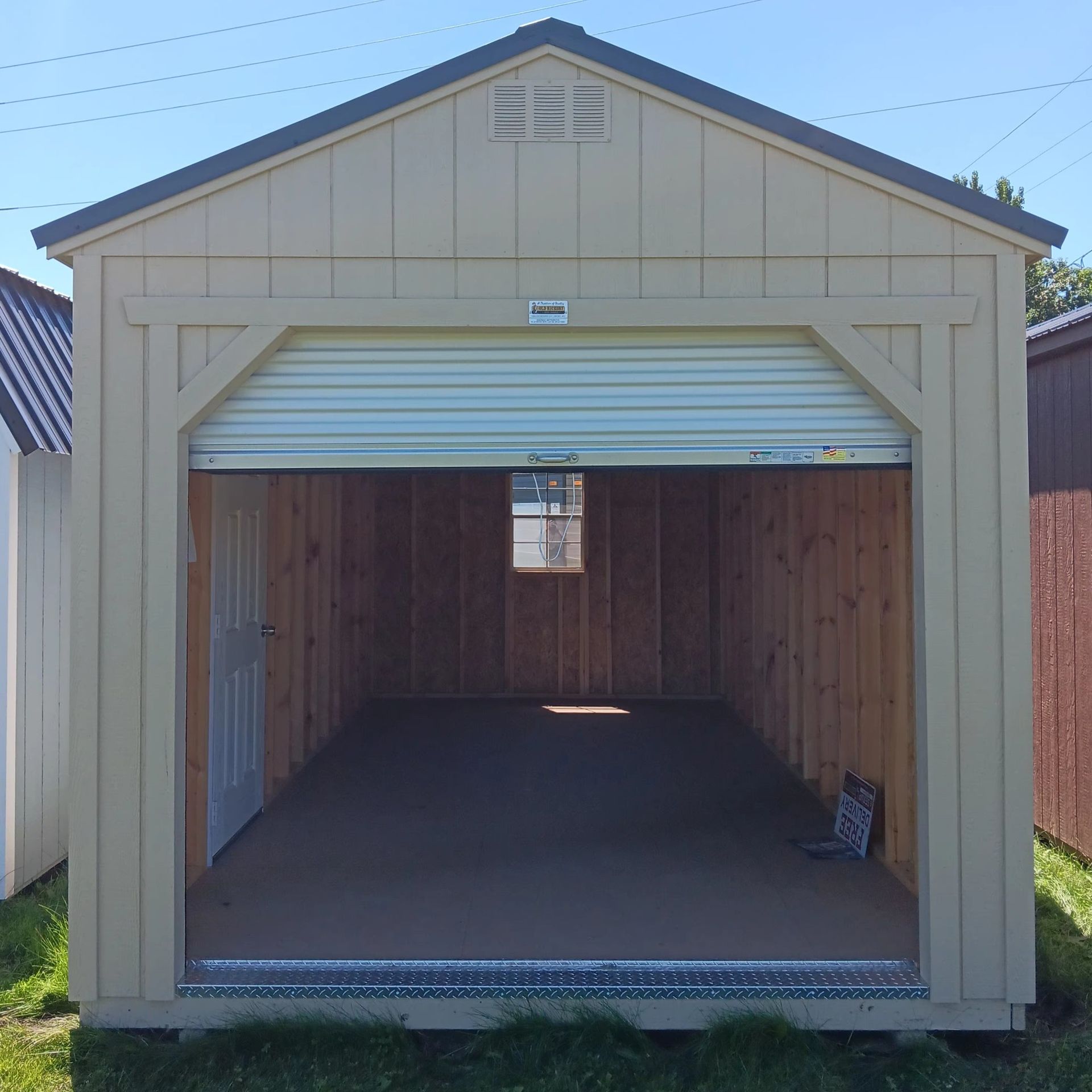 Tan shed with a roll-up garage door open. Interior wood walls and floor. Small window in the back.