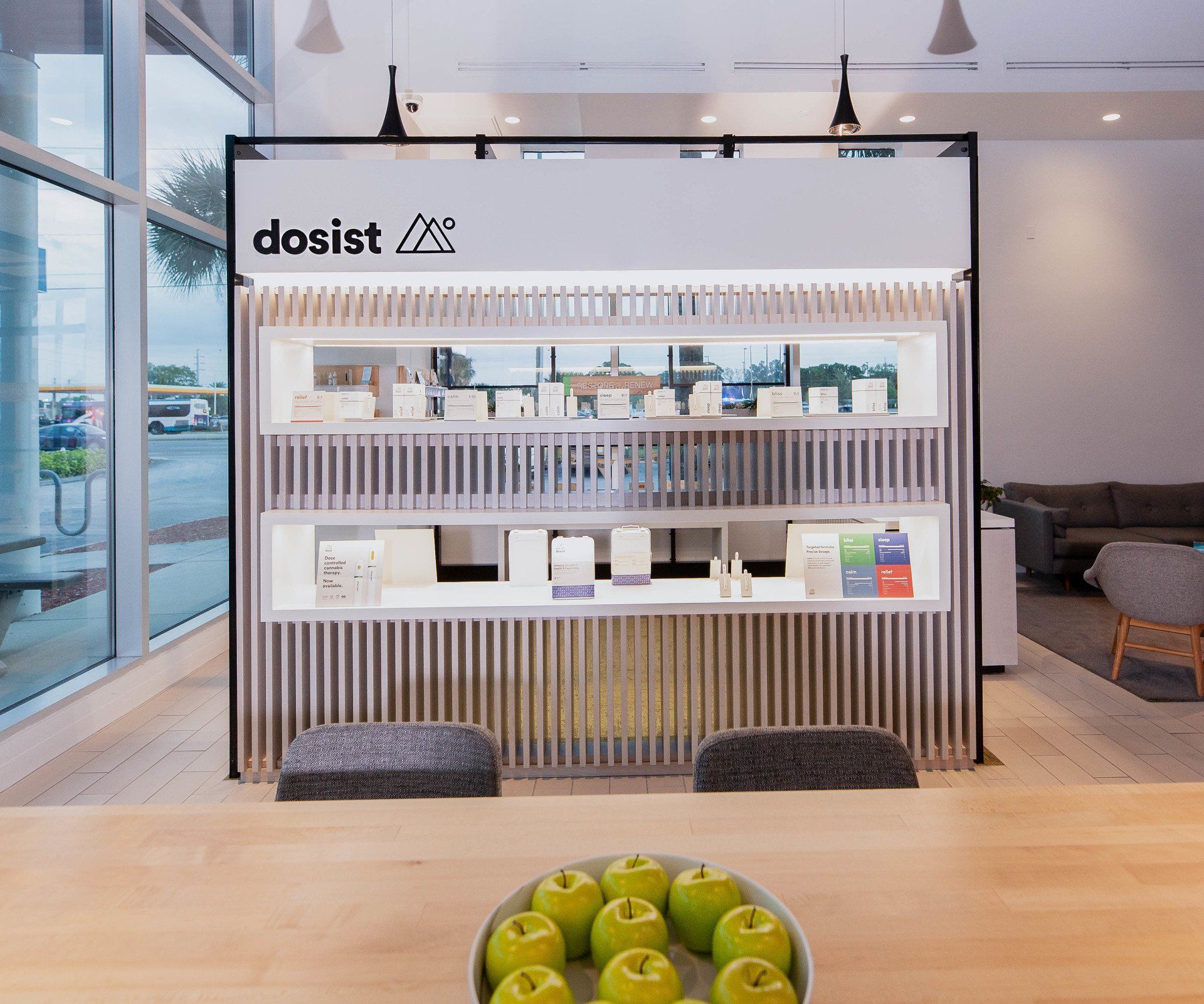 A bowl of green apples sits on a wooden table in front of a store called dosist