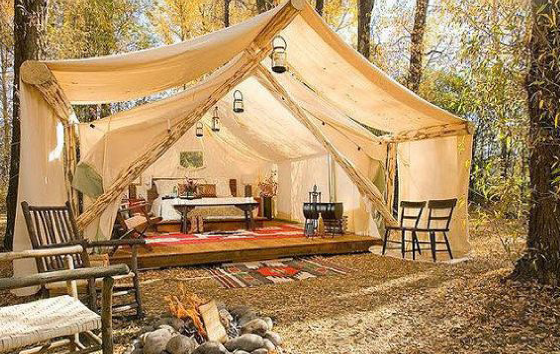 A large tent is sitting in the middle of a forest next to a fire pit.