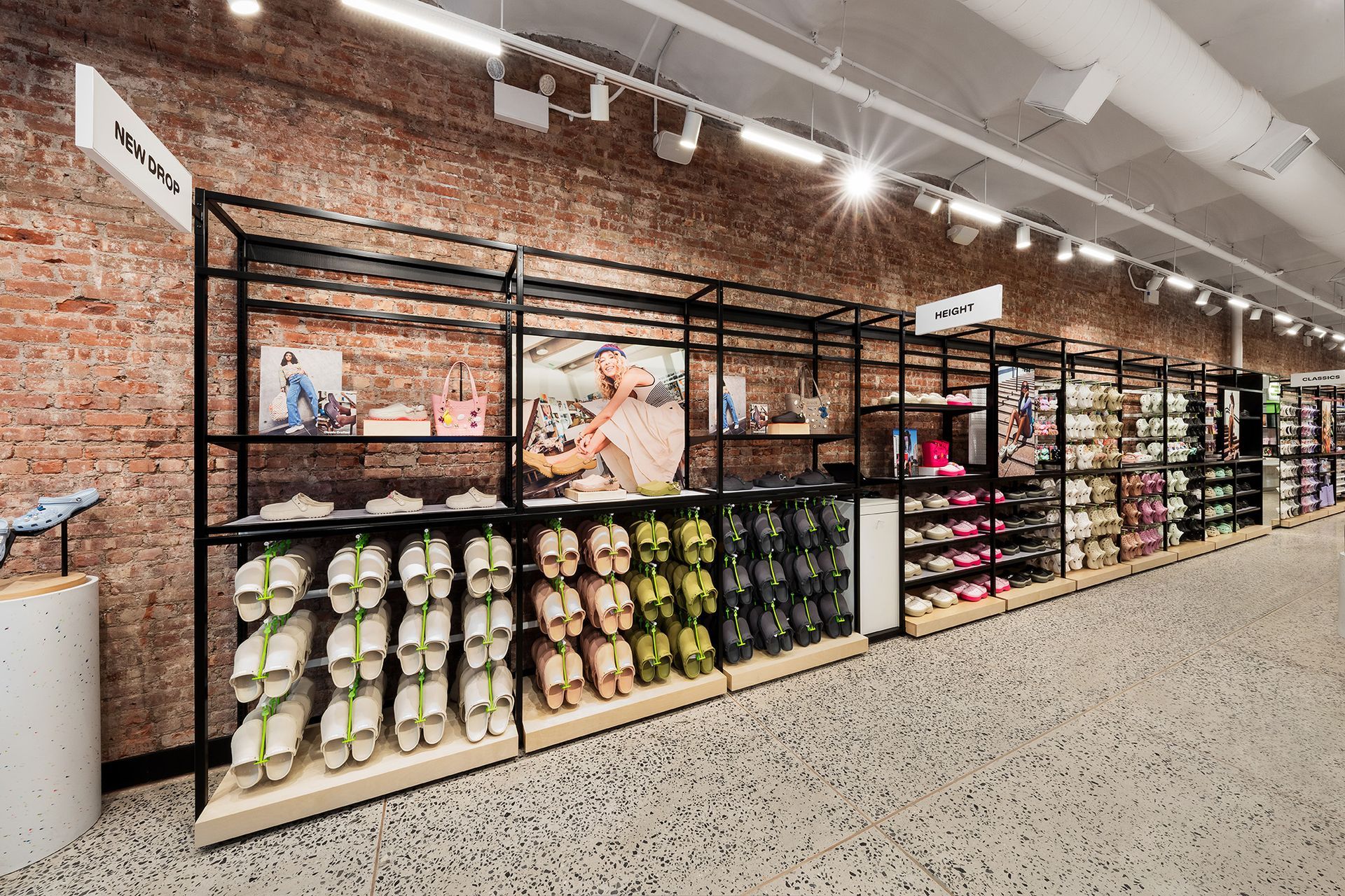Crocs hoe store interior with shelves displaying various shoes. A cashier stands near the center. Brick wall background.