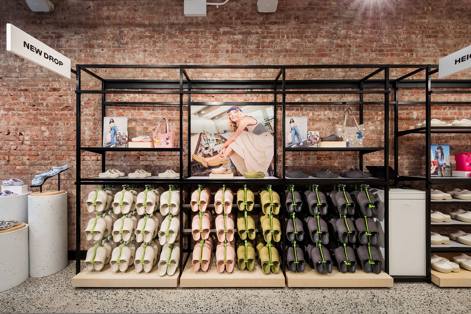Crocs Shoe display in a store with multiple colored sandals on a wooden shelf, set against a brick wall backdrop.