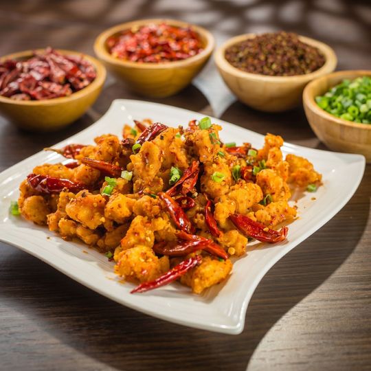 Plate of crispy fried cauliflower with red chilies, garnished with herbs, on a wooden table with spice bowls in back