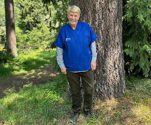Person in a blue shirt and green pants standing by a large tree in a grassy wooded area.