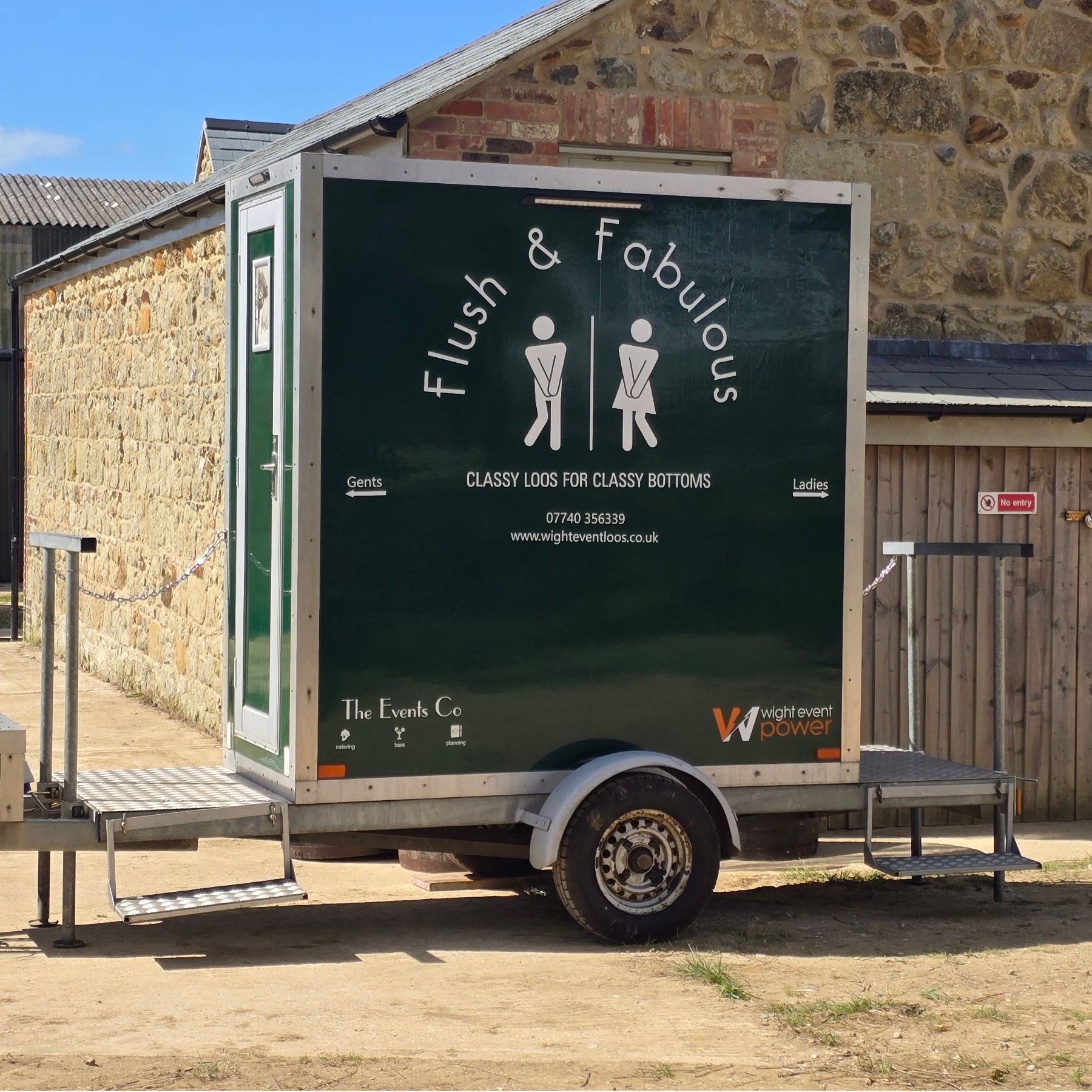 Portable toilet trailer with a “flush & follies” sign parked outdoors.