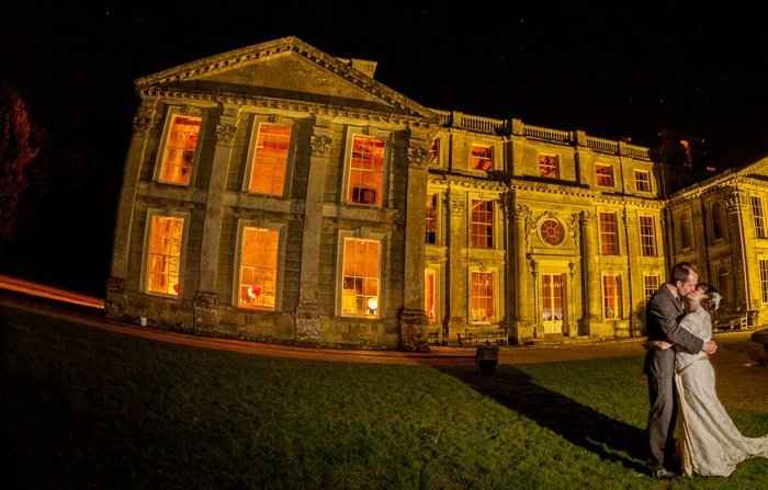 A bride and groom are kissing in front of a large building at night.