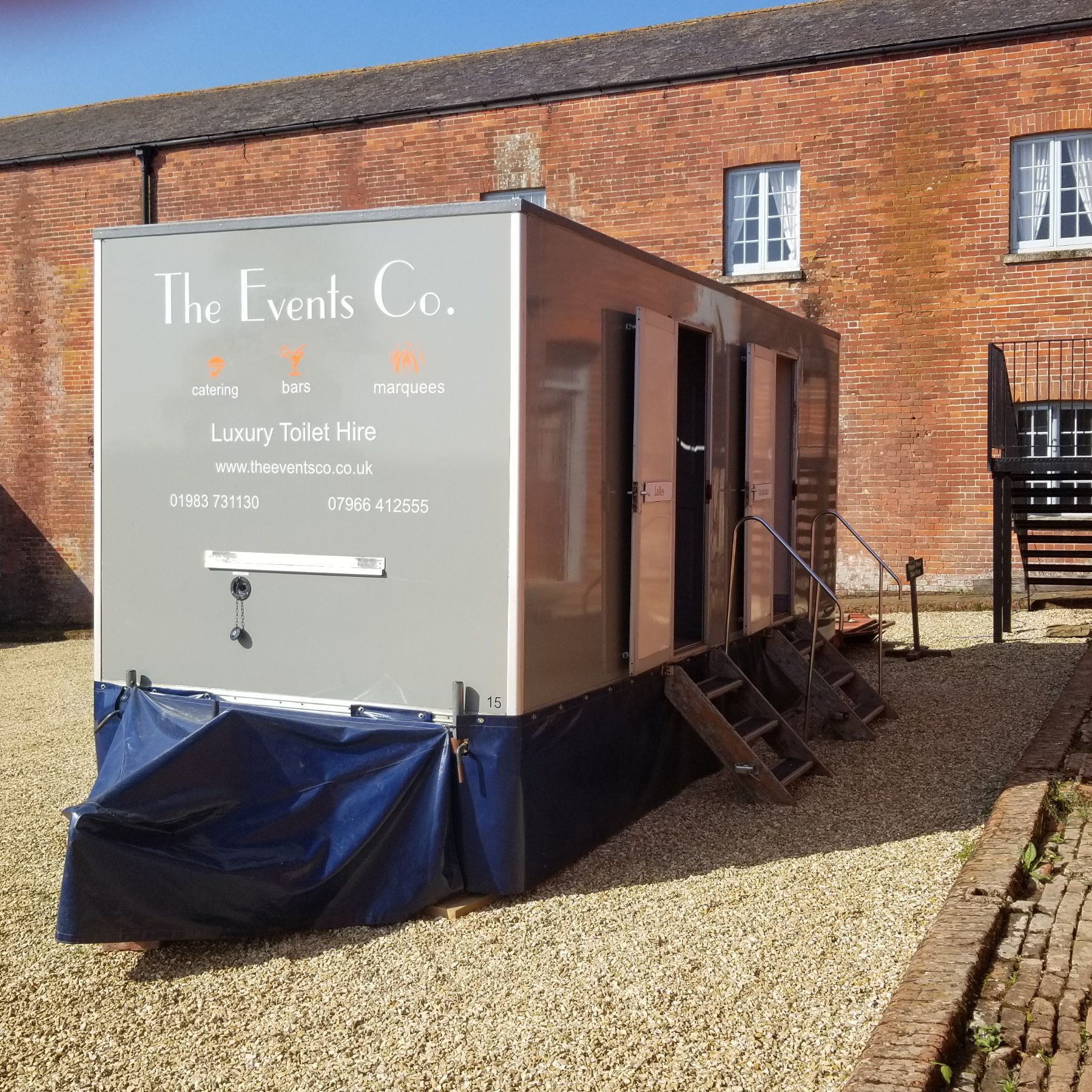 Gray portable toilet unit with “The Lovells Co.” branding parked beside a brick building on gravel