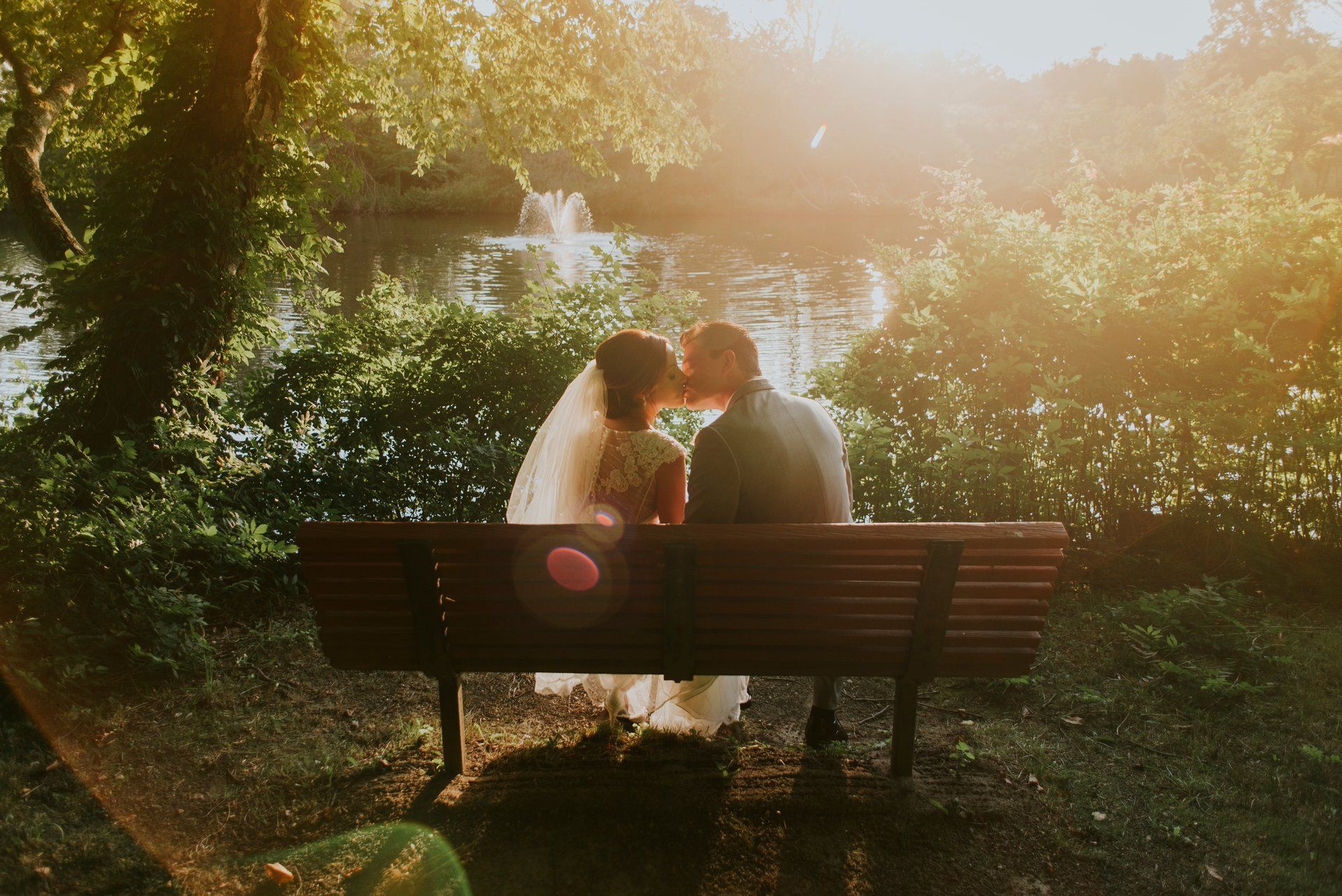 A bride and groom are kissing on a park bench.