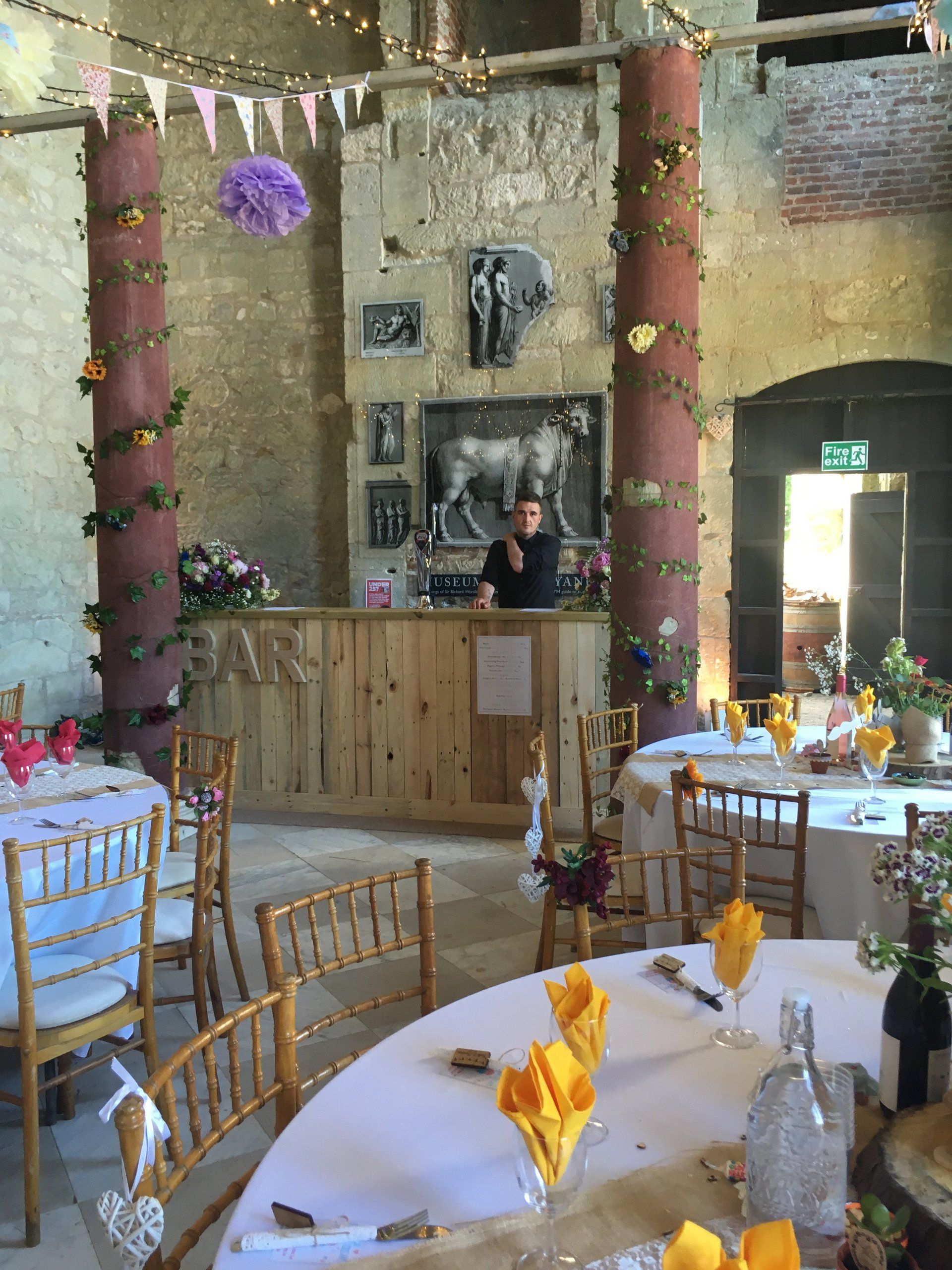 A man is standing behind a bar in a room with tables and chairs.