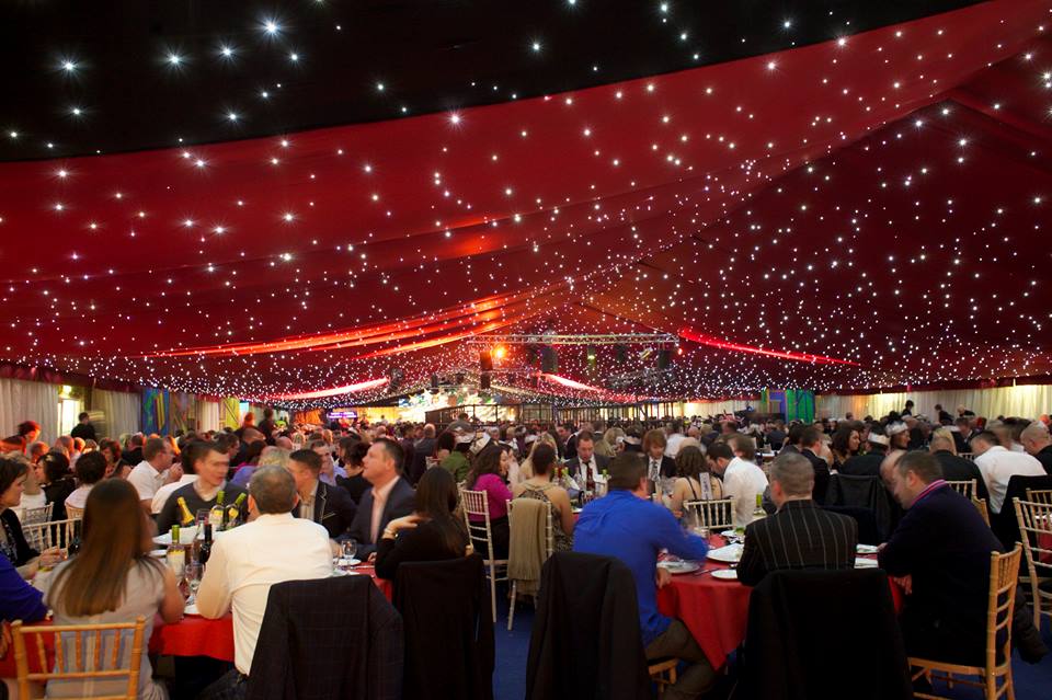A large group of people are sitting at tables under a red tent.