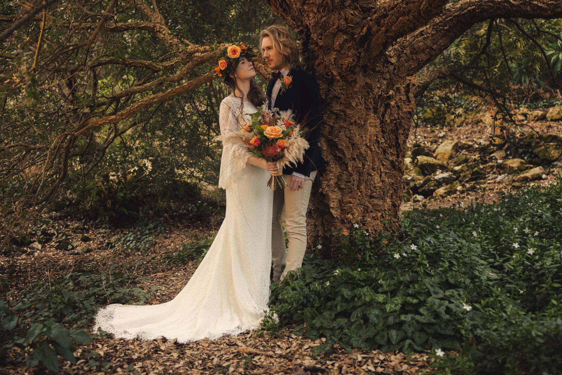 A bride and groom are standing next to a tree in the woods.