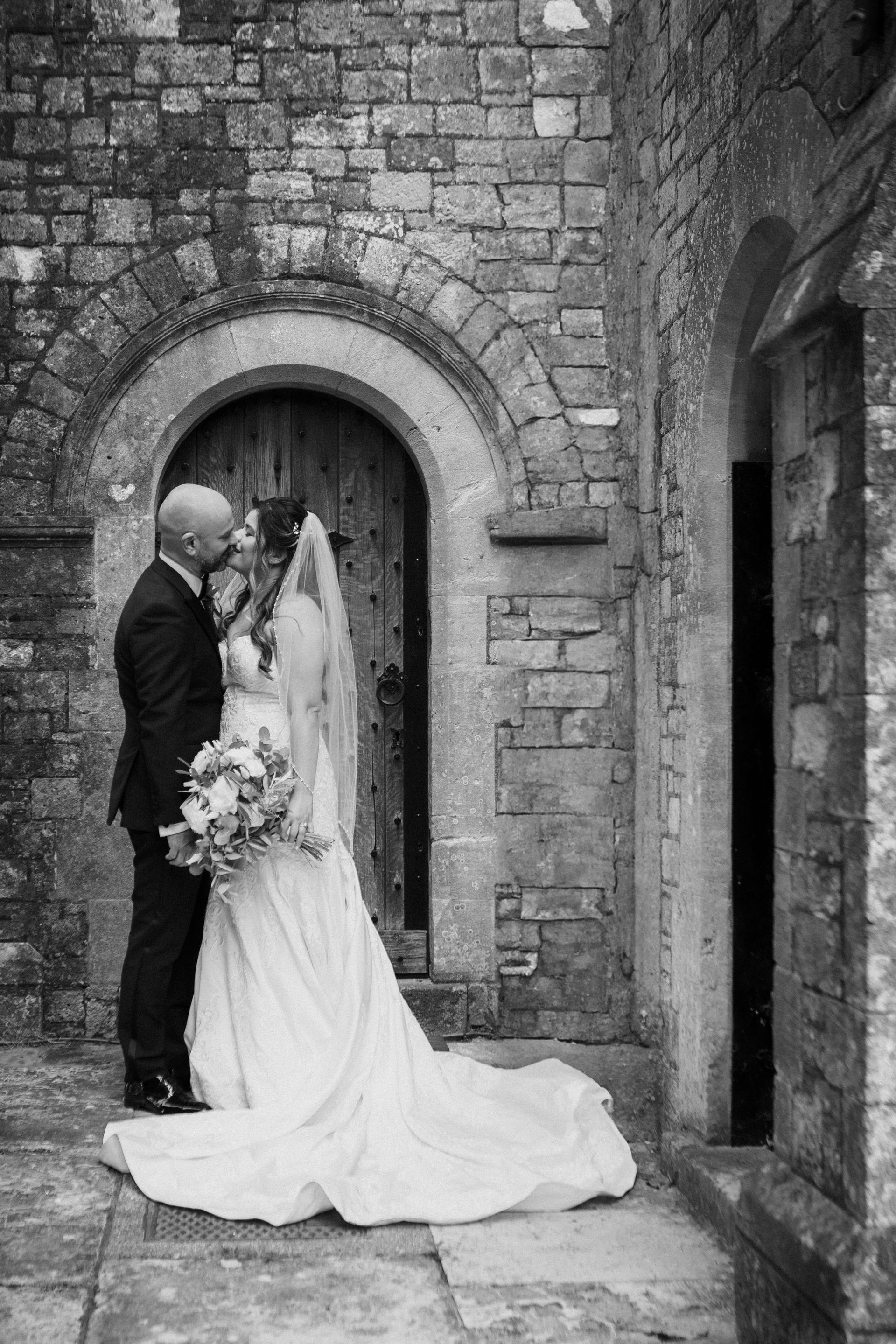 A black and white photo of a bride and groom kissing in front of a brick building.