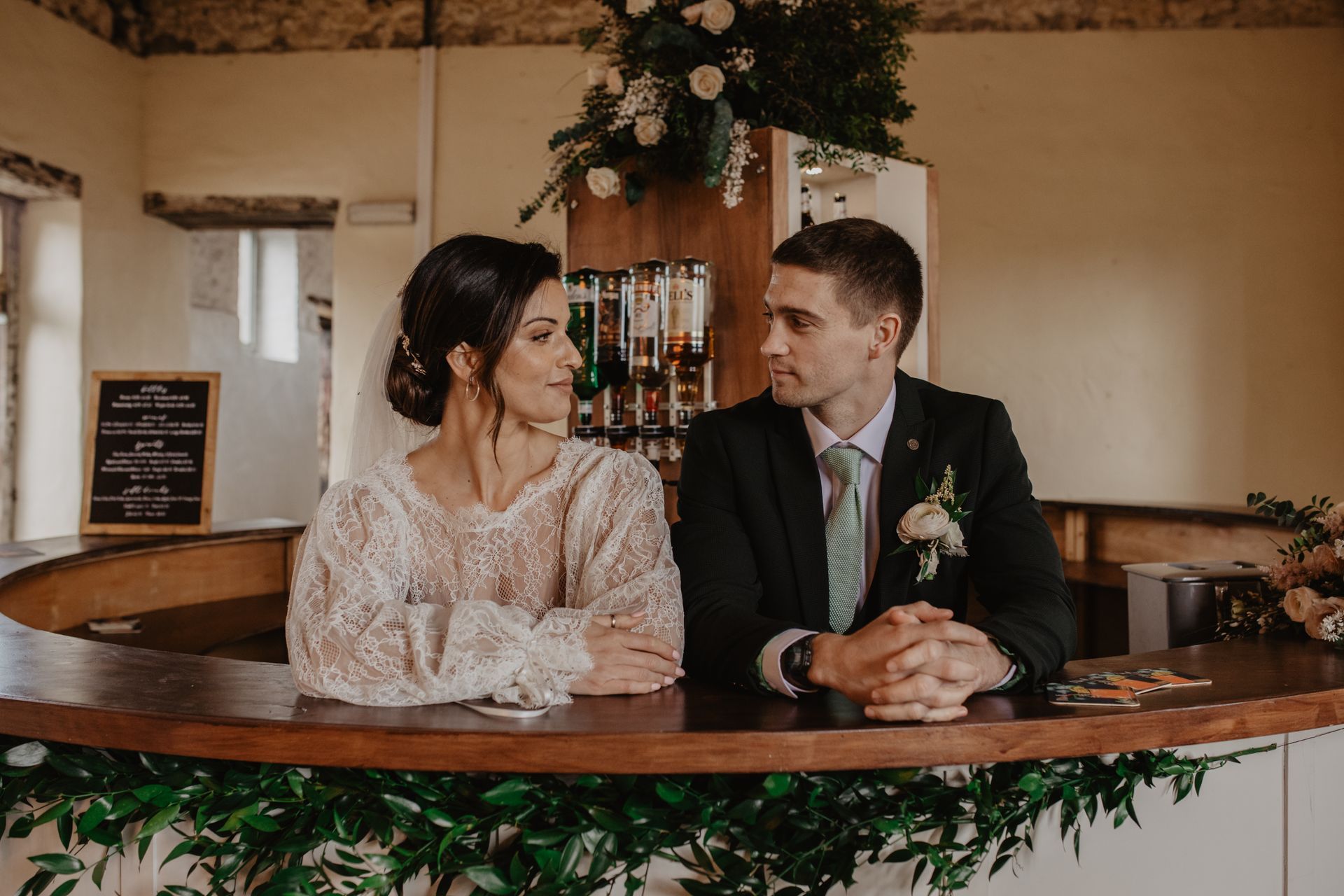 A bride and groom are sitting at a bar looking at each other.