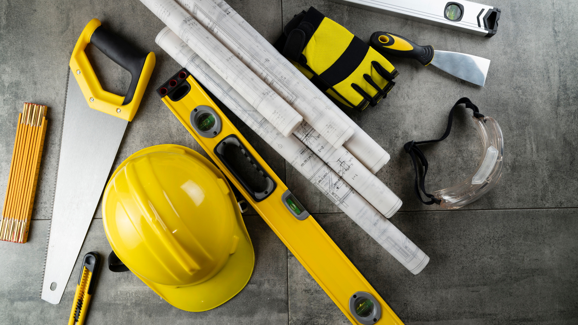 A hard hat , saw , level , gloves , and blueprints are on a table.