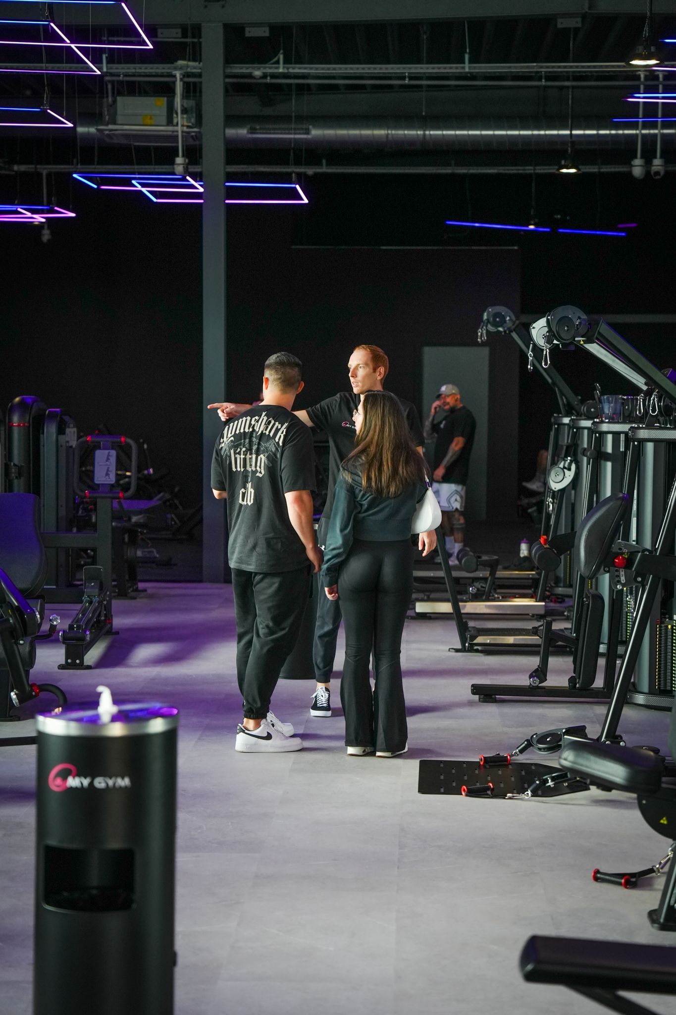 Mensen in een sportschool. Iemand wijst naar de apparaten. Paars-witte verlichting, donker interieur.