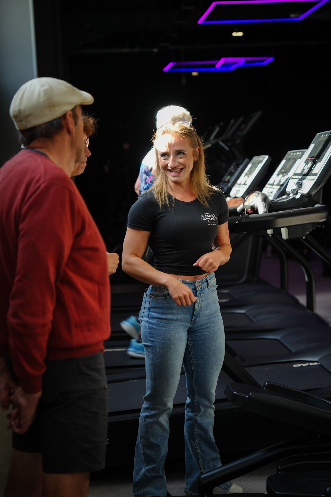 Een vrouw in spijkerbroek en zwart shirt lacht terwijl ze praat met een man in een rood shirt in een sportschool met loopbanden.
