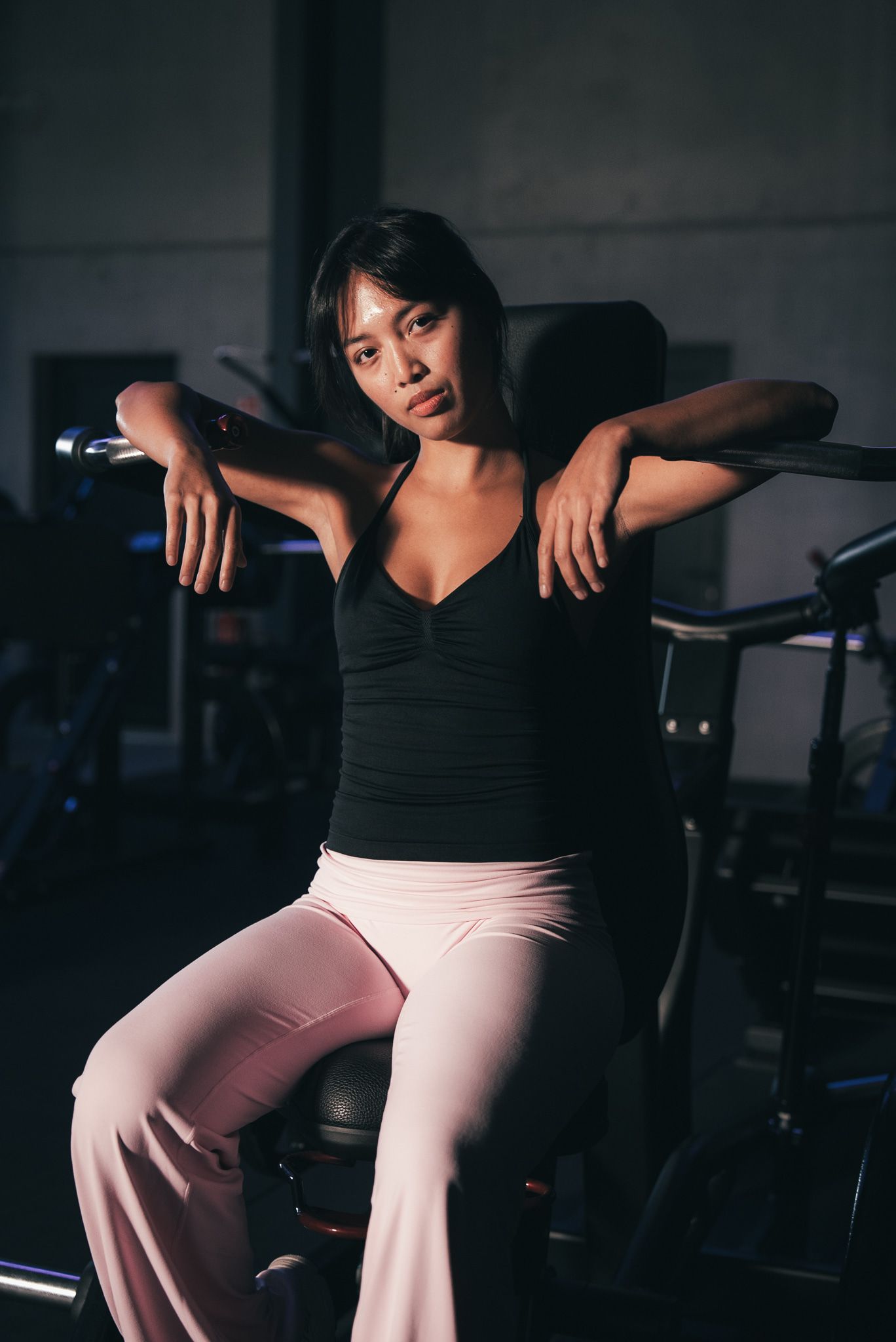 Vrouw rust op fitnessapparatuur, gekleed in een zwart tanktopje en een roze broek.