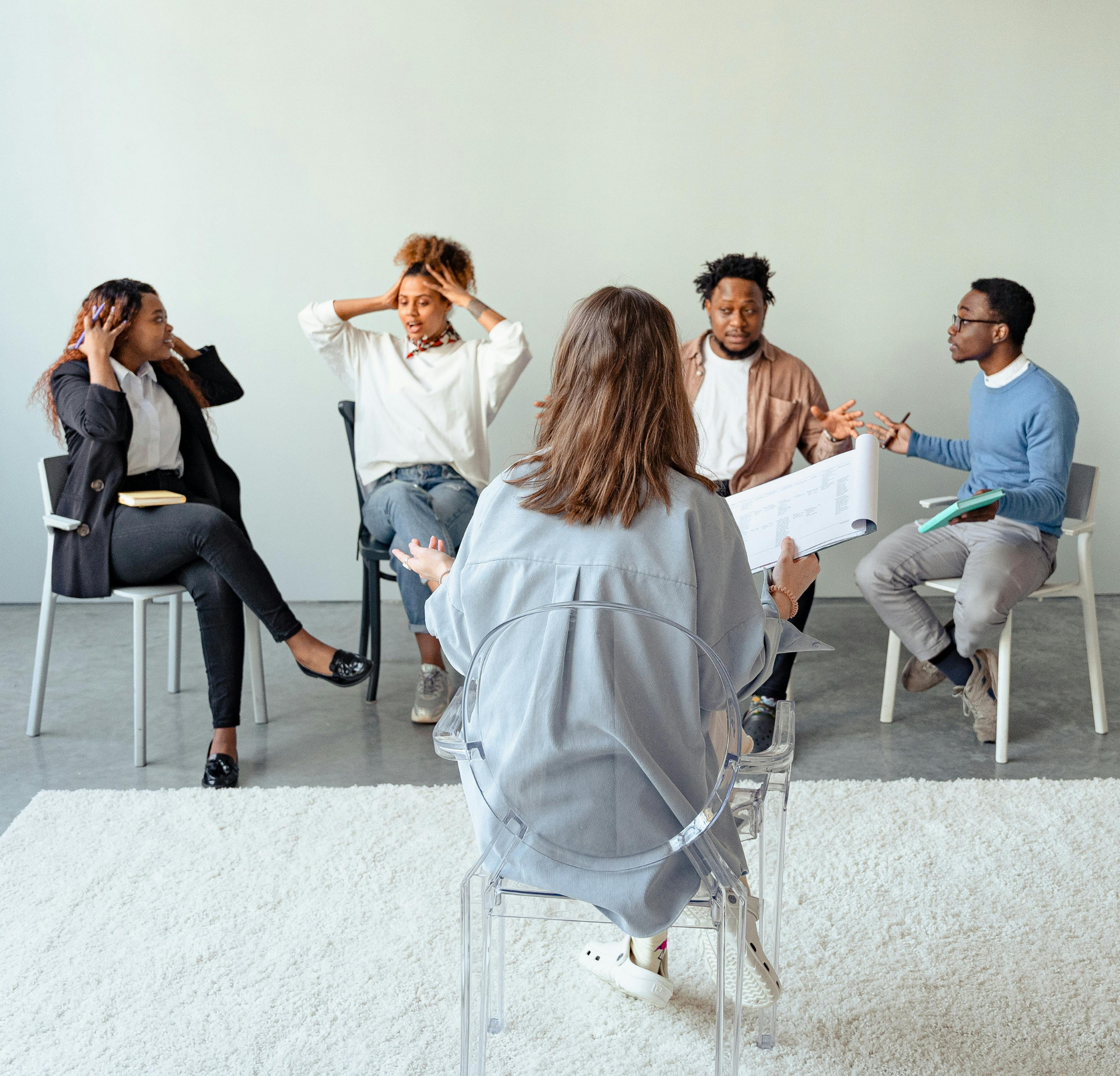 Five people sit in a circle in a bright room, participating in a group meeting or therapy session.