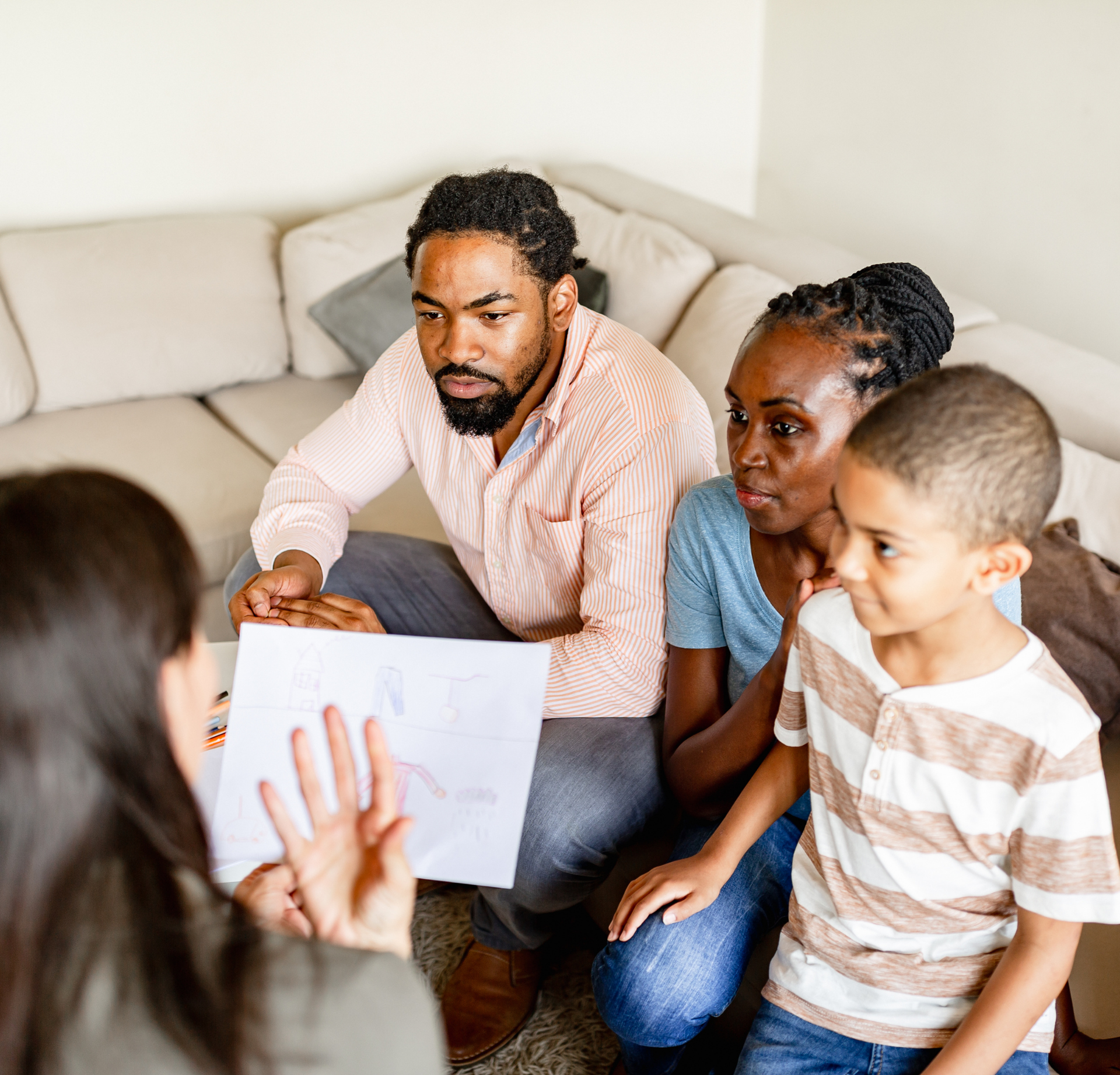 A person holds a document while explaining it to two people sitting on a couch in a brightly lit room.