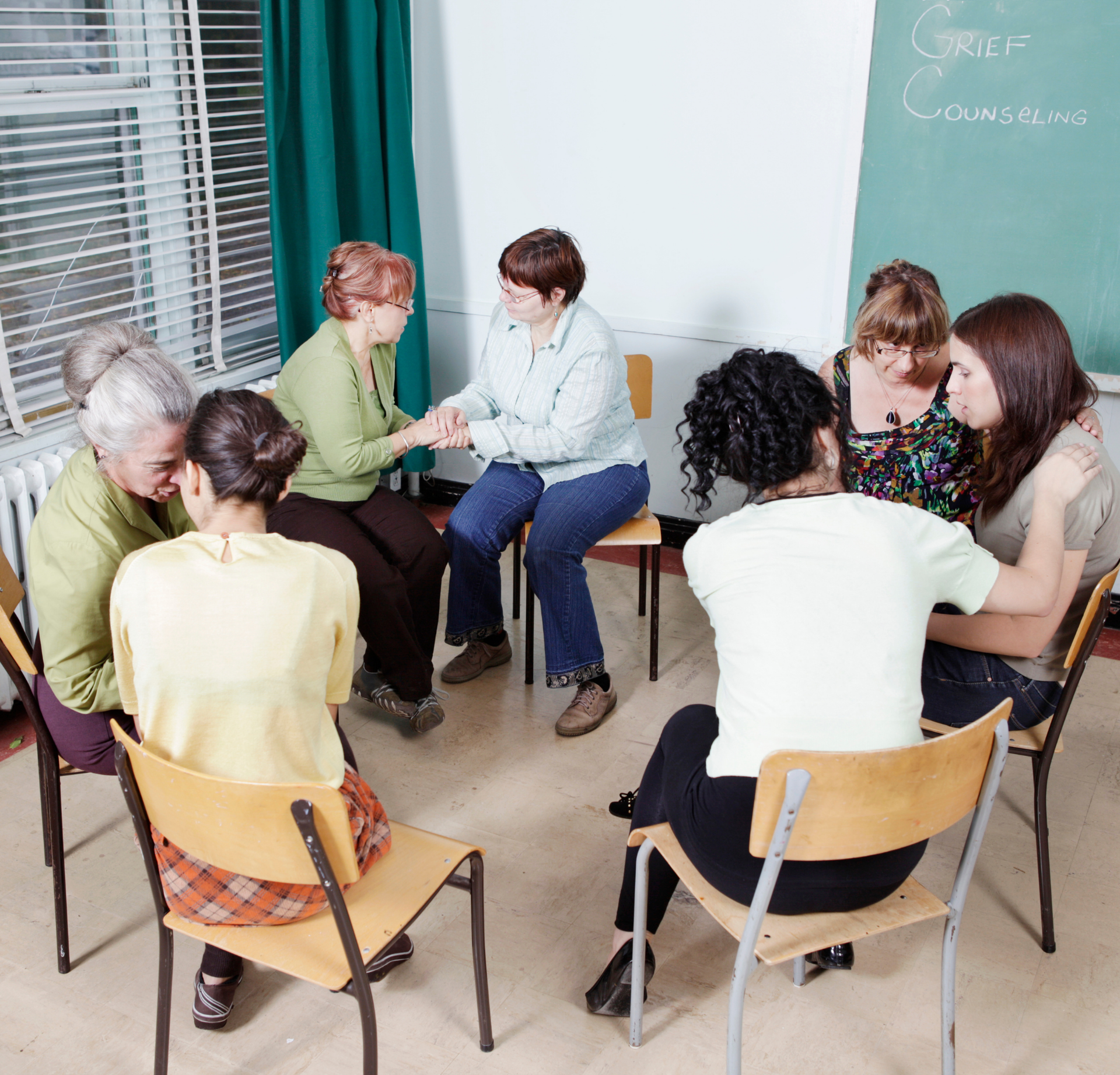 A group sits in a circle at a support meeting, with some holding hands or hugging.