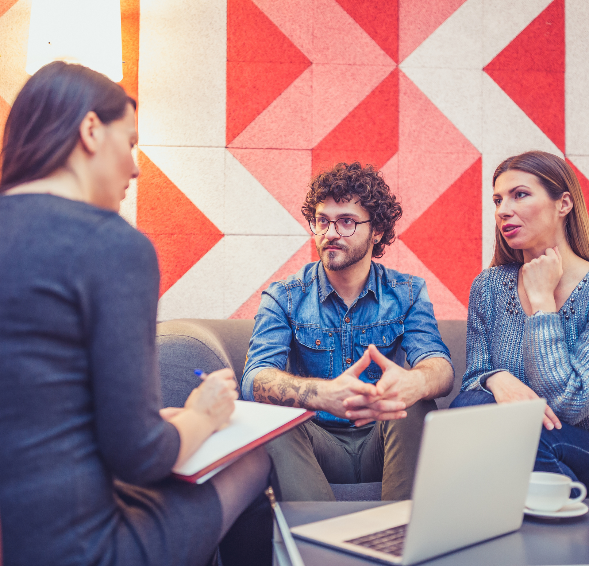 A person takes notes while two others sit on a couch in a modern office with a red and white geometric wall pattern.
