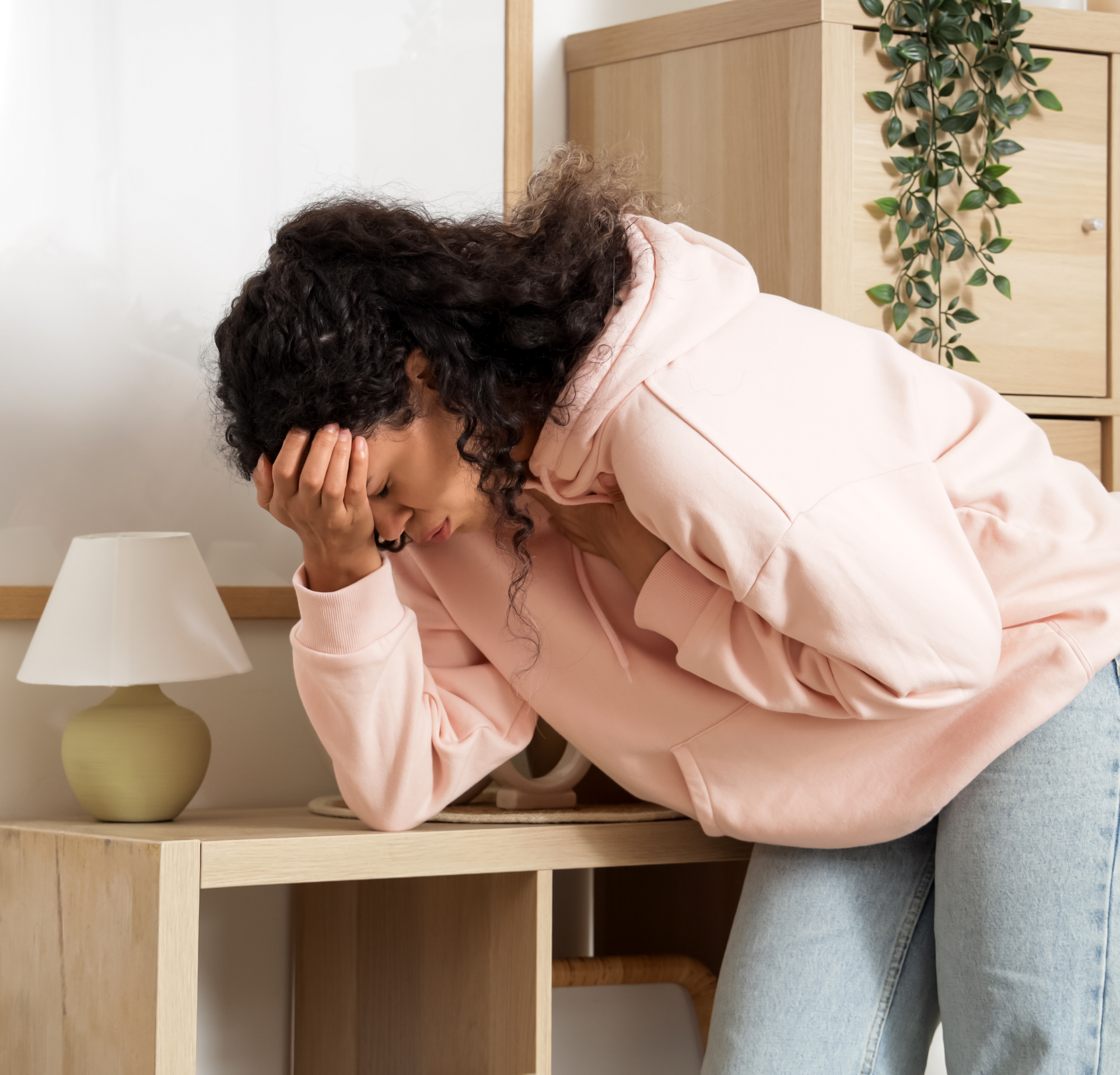 A person in a light pink hoodie leaning over a wooden desk with their head in their hand, looking distressed.