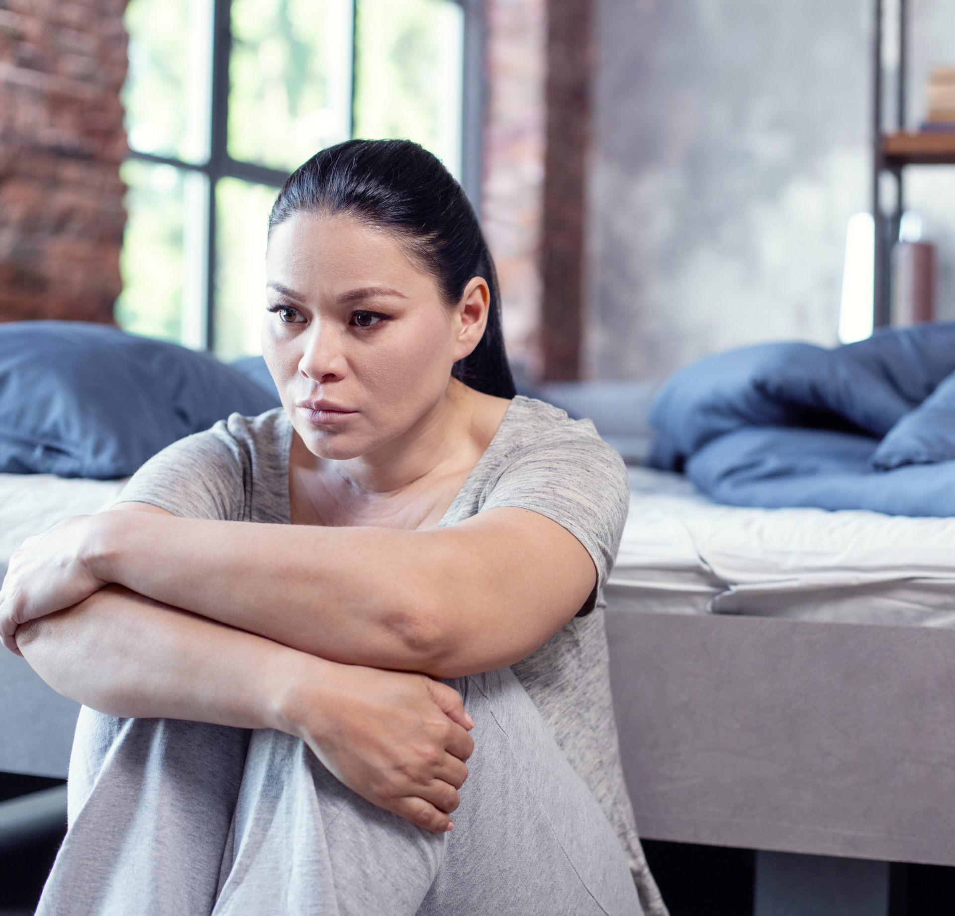 A person sits on the floor by a bed, holding their knees with a somber expression in a bedroom.