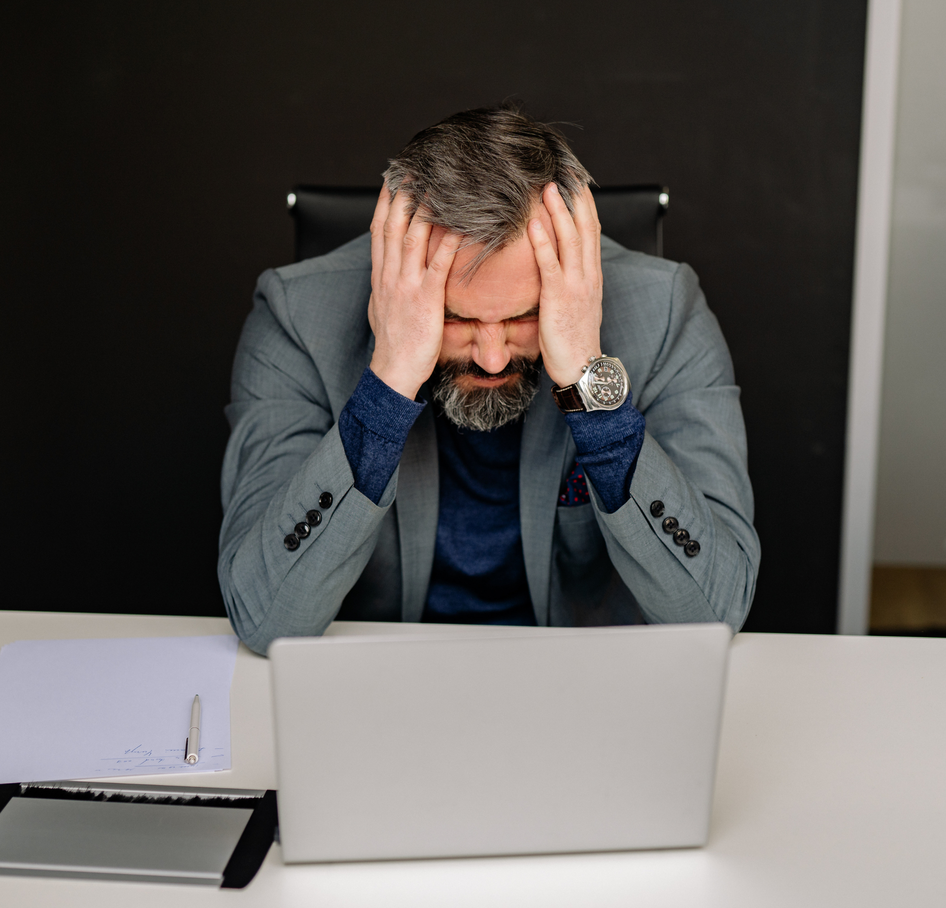 A person with a gray beard in a suit sits at a desk with a laptop, head in their hands, looking stressed.