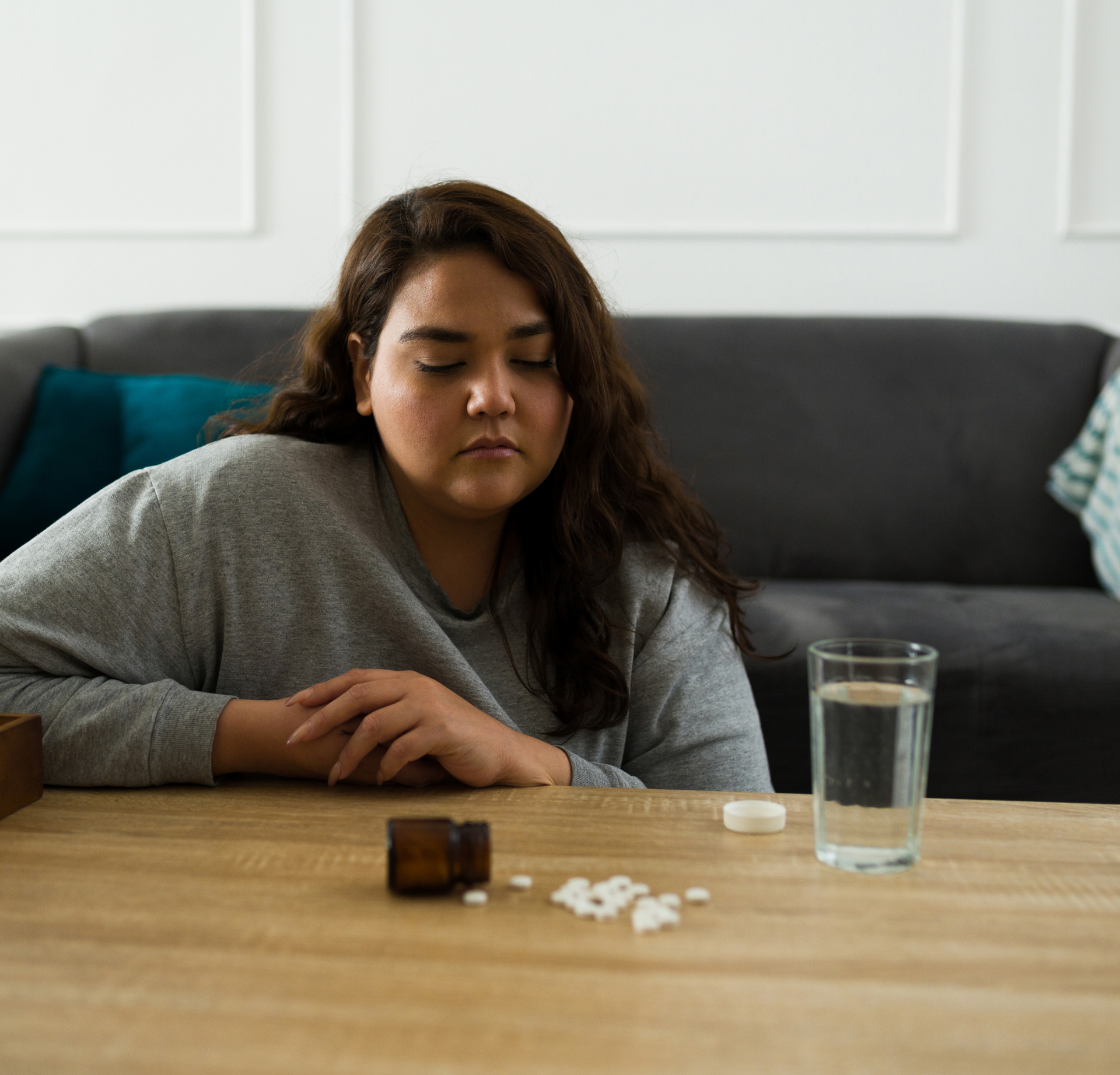 A person sits at a wooden table with a glass of water and several loose white pills spilled from an open bottle.