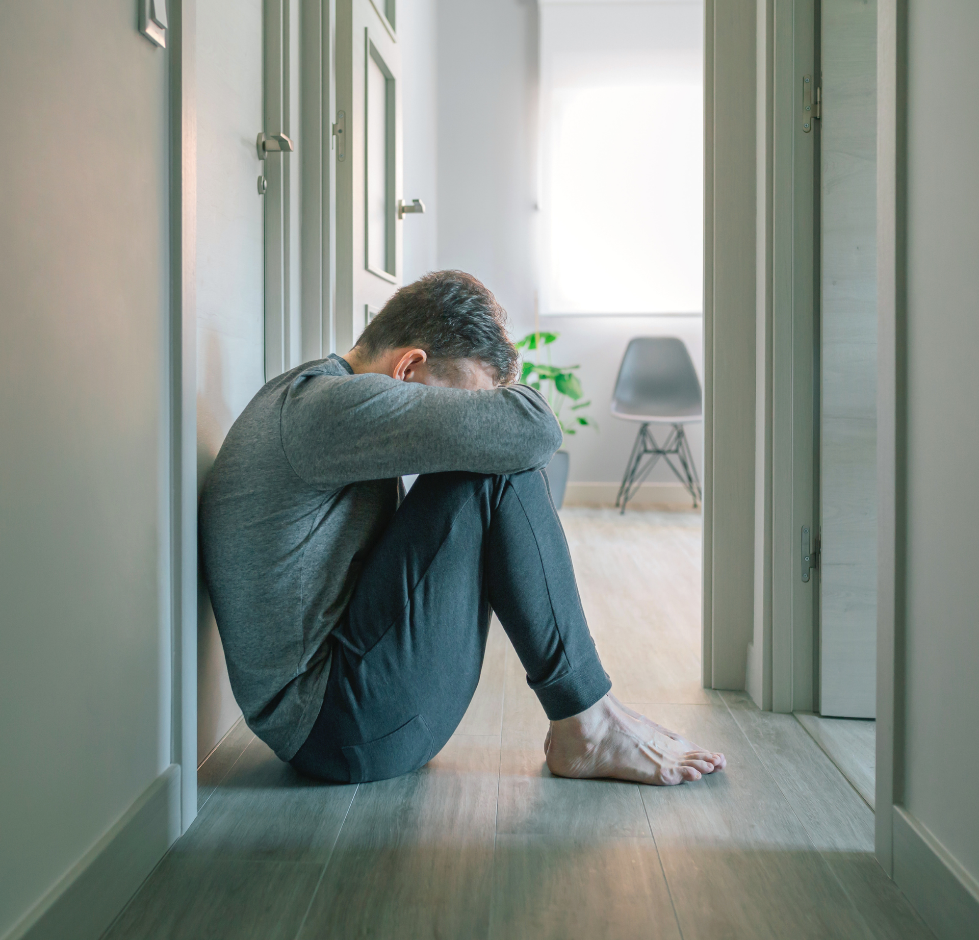 A person sitting on a hallway floor, hugging their knees with their head bowed in a posture of distress.