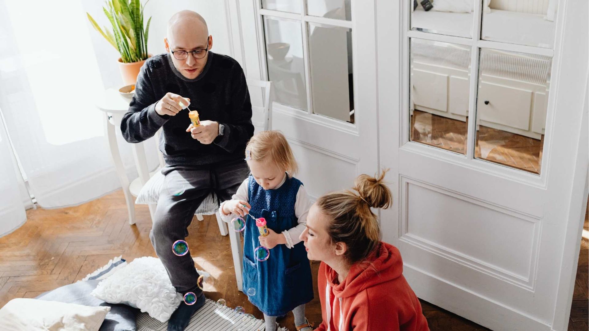 Family indoors blowing bubbles. Man sitting, child and woman standing, all smiling.