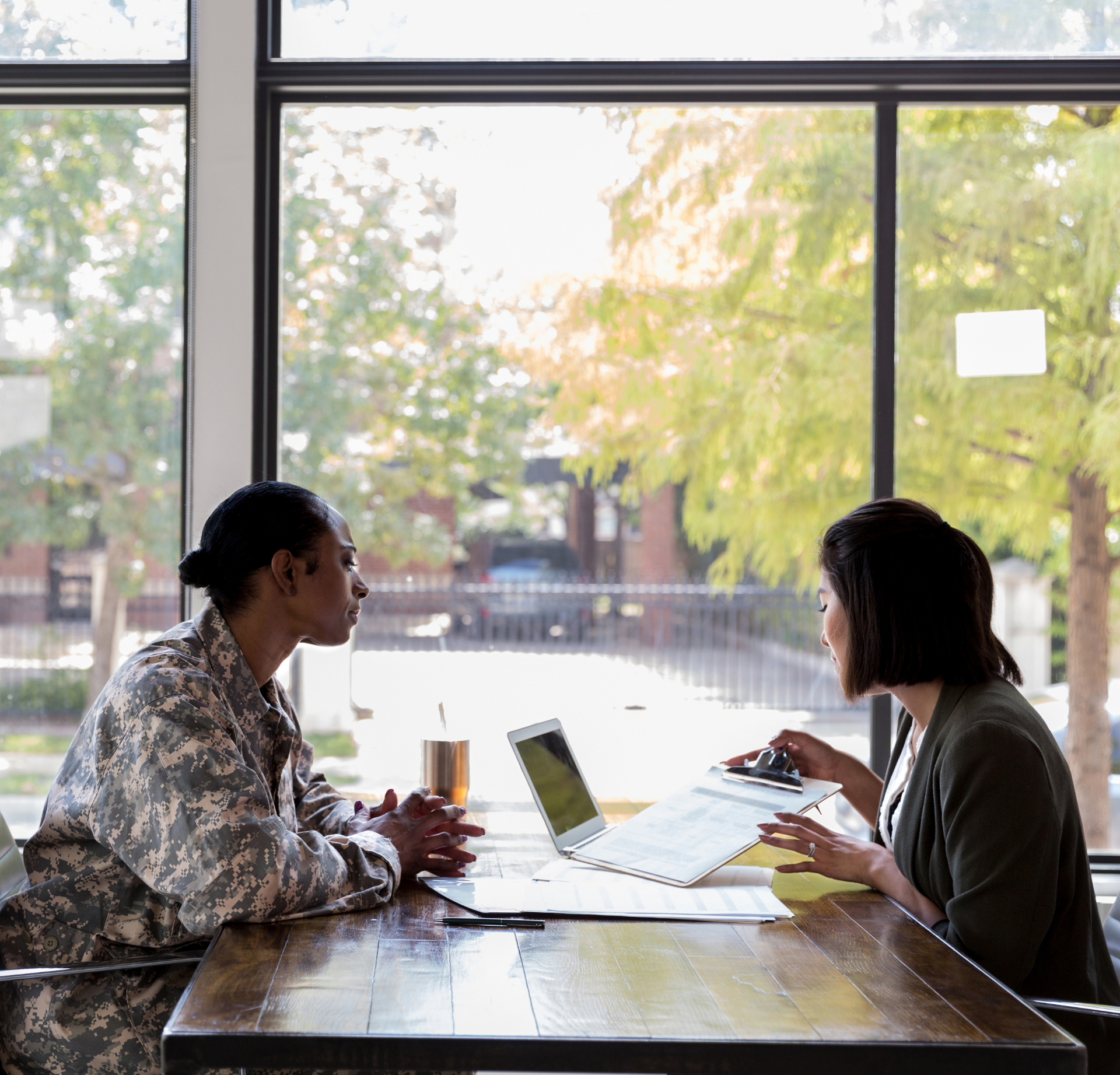A person in military uniform sits at a table with an advisor in a professional setting, discussing documents and a laptop.