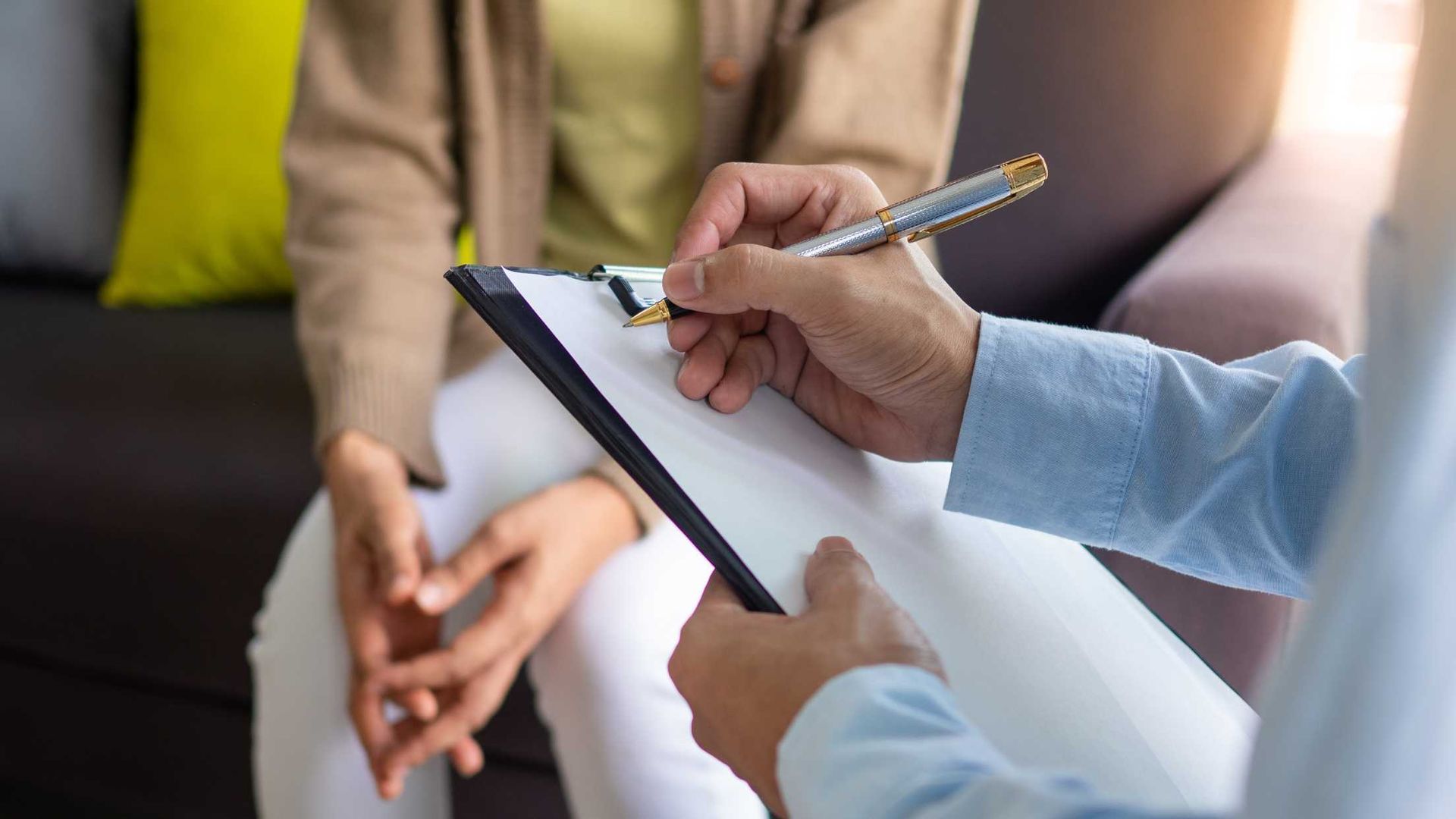 Person taking notes on clipboard; another person seated.