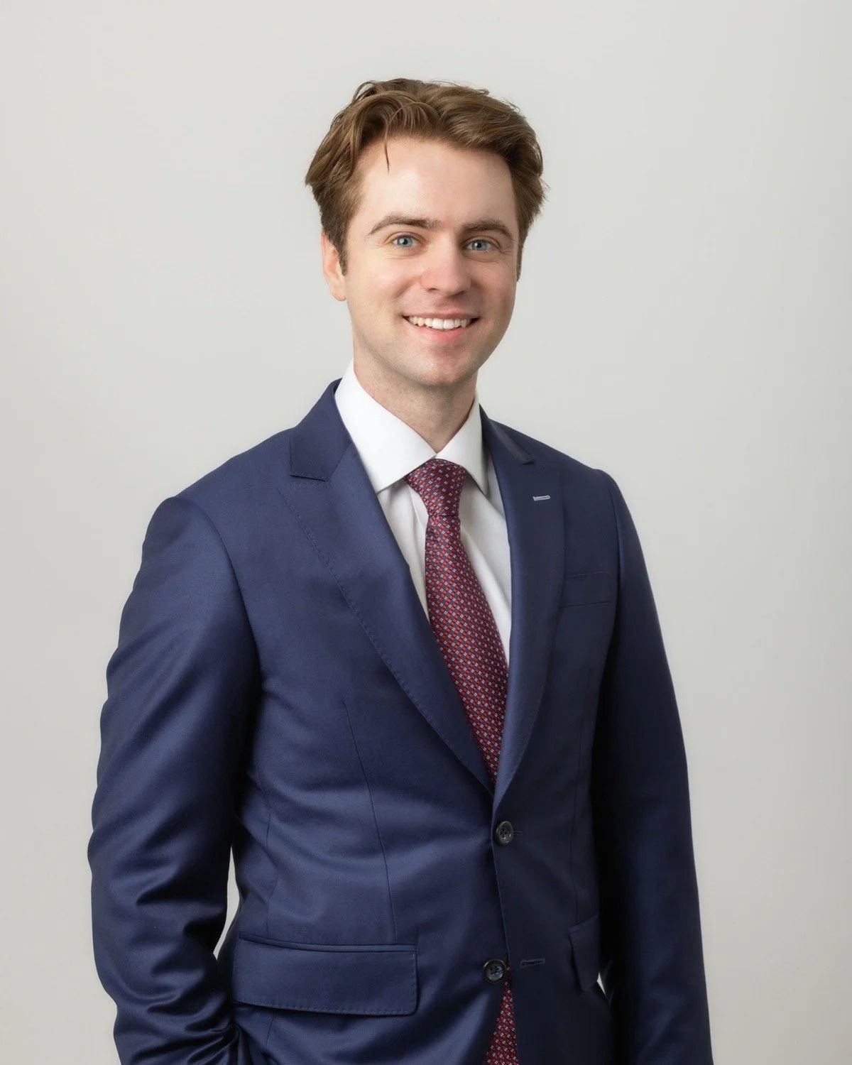 Man in navy suit and maroon tie smiles, posing against a white background.