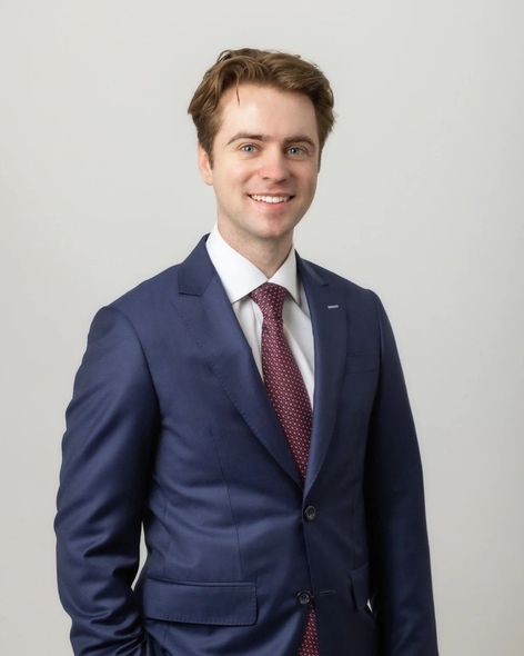 Man in navy suit and maroon tie smiles, posing against a white background.