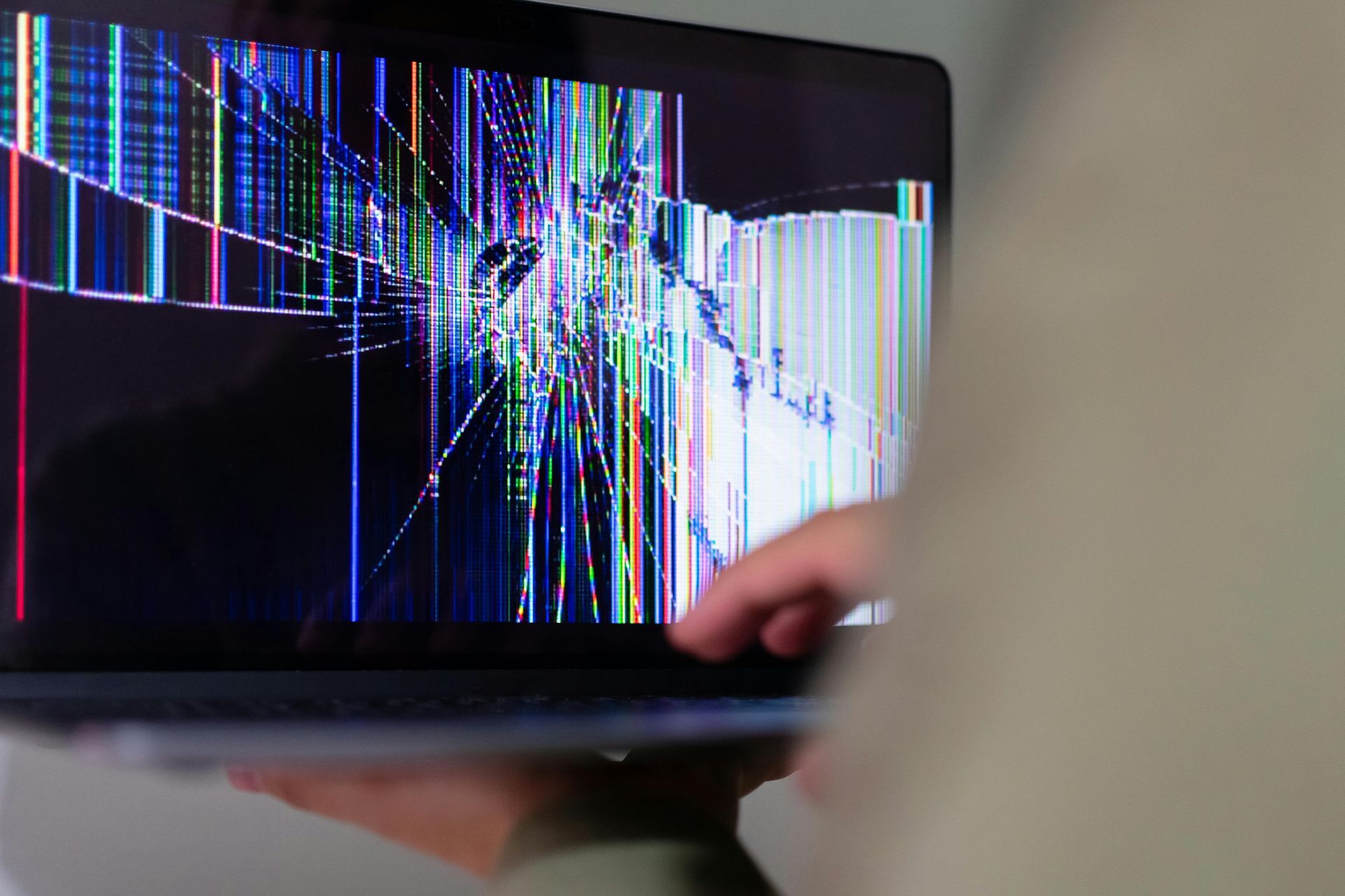 Person holding a laptop with a severely cracked screen displaying colorful, distorted lines.