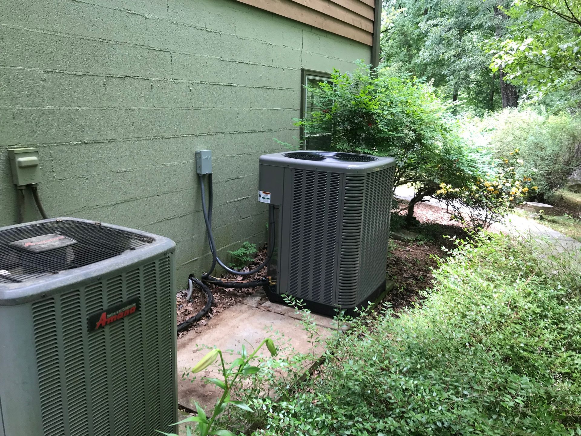 Two air conditioners are sitting next to each other on the side of a house.