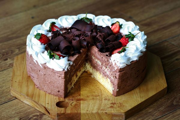 A chocolate cake with whipped cream and strawberries on top is on a wooden cutting board.