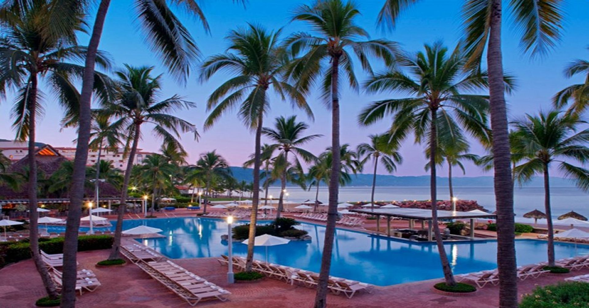 A large swimming pool surrounded by palm trees next to the ocean. Vacation Travel Exclusives