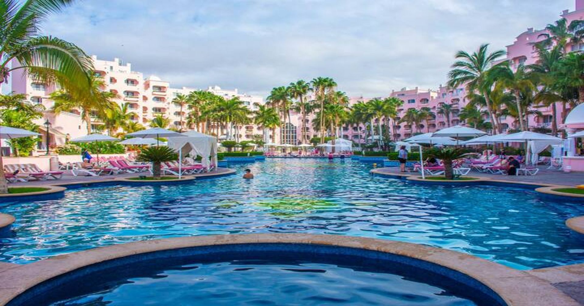 A large swimming pool surrounded by palm trees and buildings at a resort. Vacation Travel Exclusives