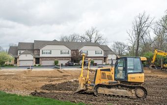 A yellow bulldozer clears dirt on a construction site in front of a multi-unit townhouse complex on a cloudy day.